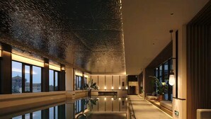 Interior moderno de un hotel con un largo pasillo, ventanas amplias, una zona de piscina y vista a un edificio tradicional.