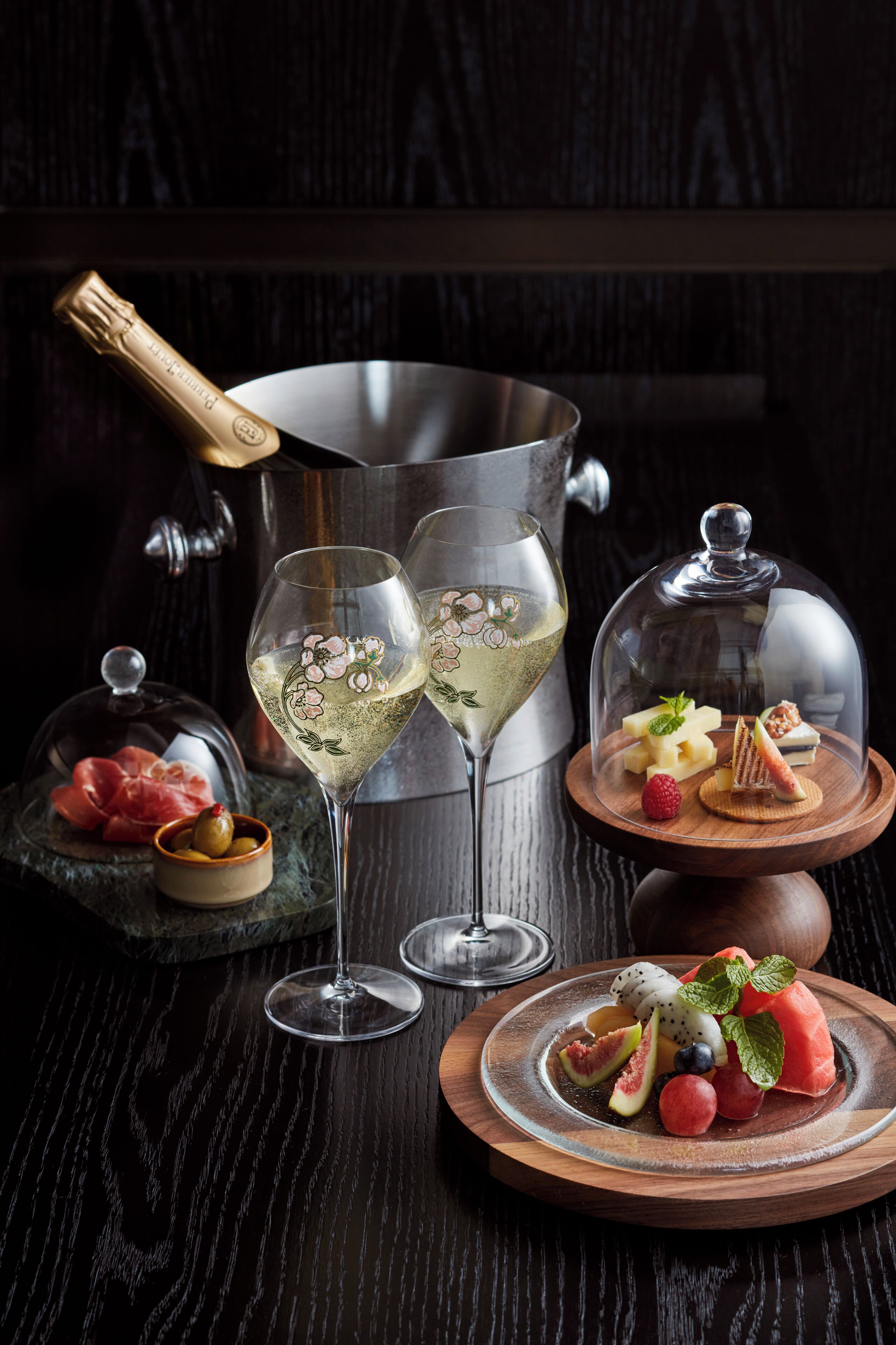 Una mesa dispuesta con dos copas de vino blanco, una botella de champán, una tabla de madera con fruta y queso, y una campana de cristal cubriendo un postre.