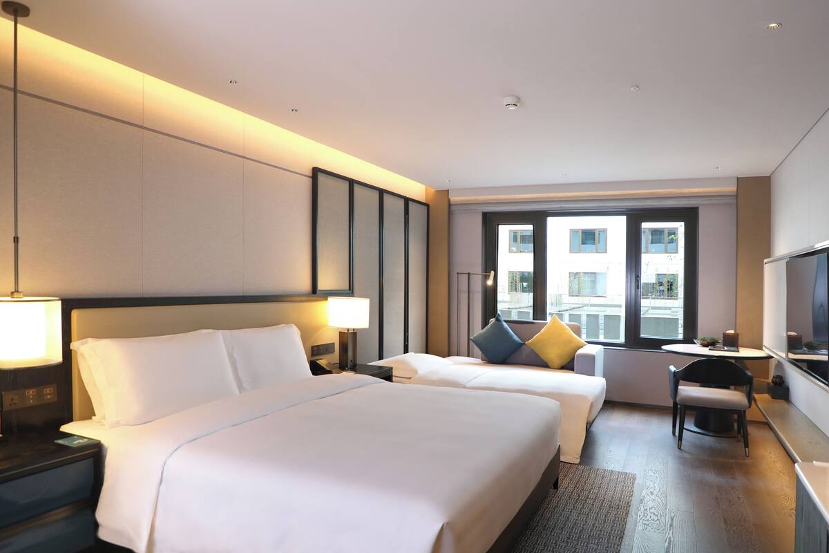 A hotel room with two beds, a desk, a chair, a TV, and a window with a city view.