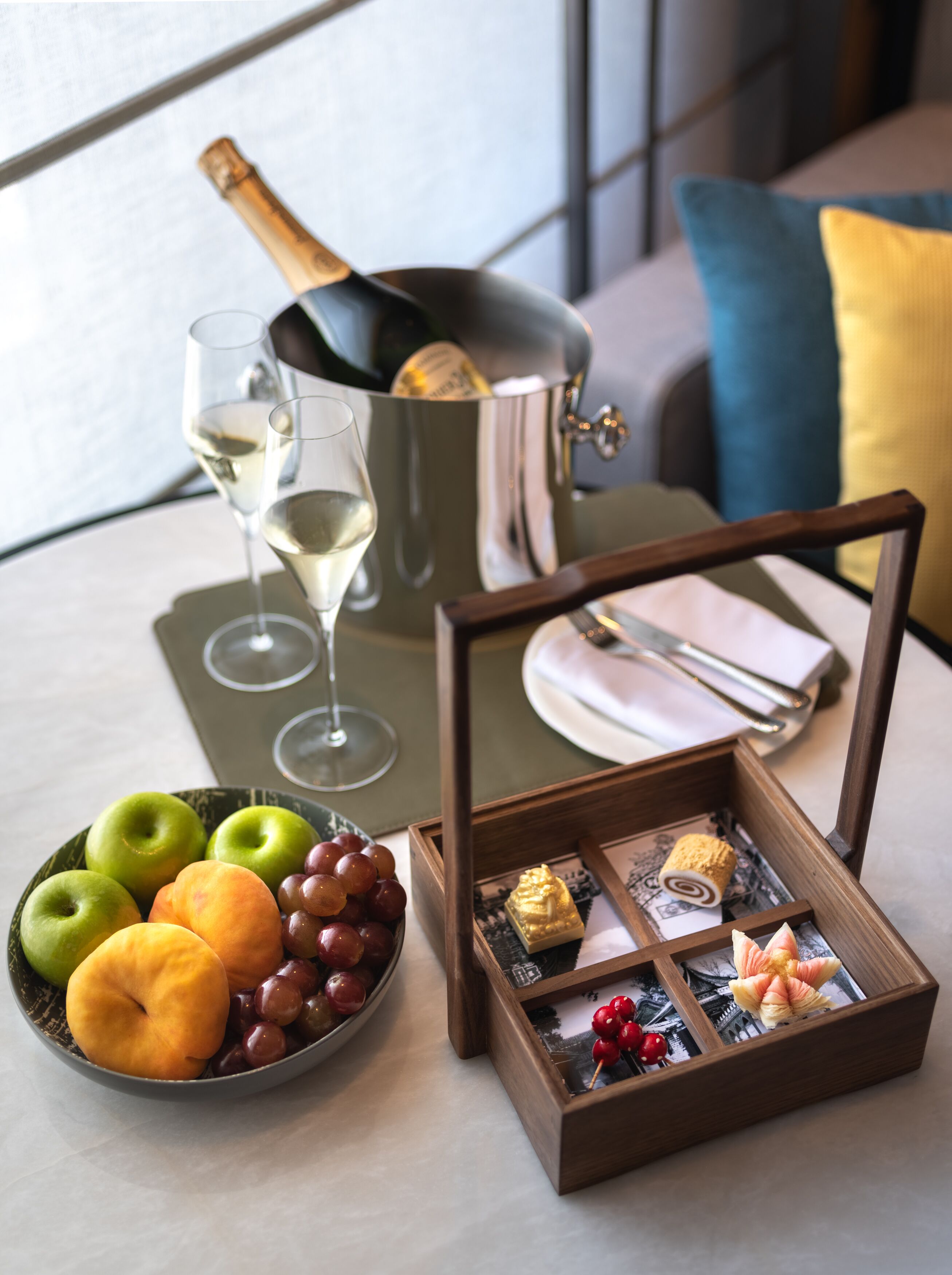A table with a bottle of champagne, two glasses of white wine, a bowl of fruit, and a wooden box containing chocolates and a decorative item.