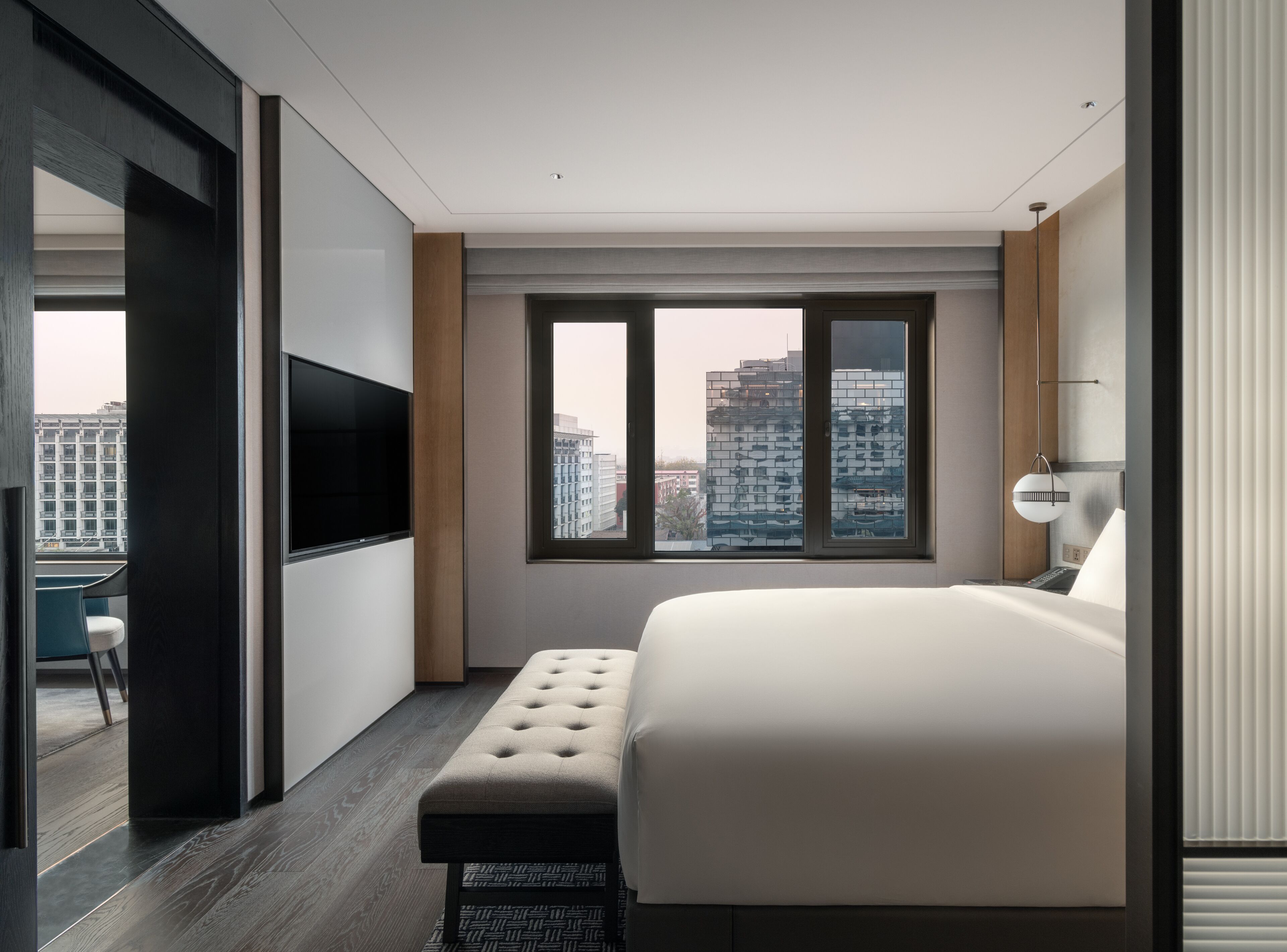 A modern bedroom with a large bed, a TV mounted on the wall, and a view of city buildings through the windows.