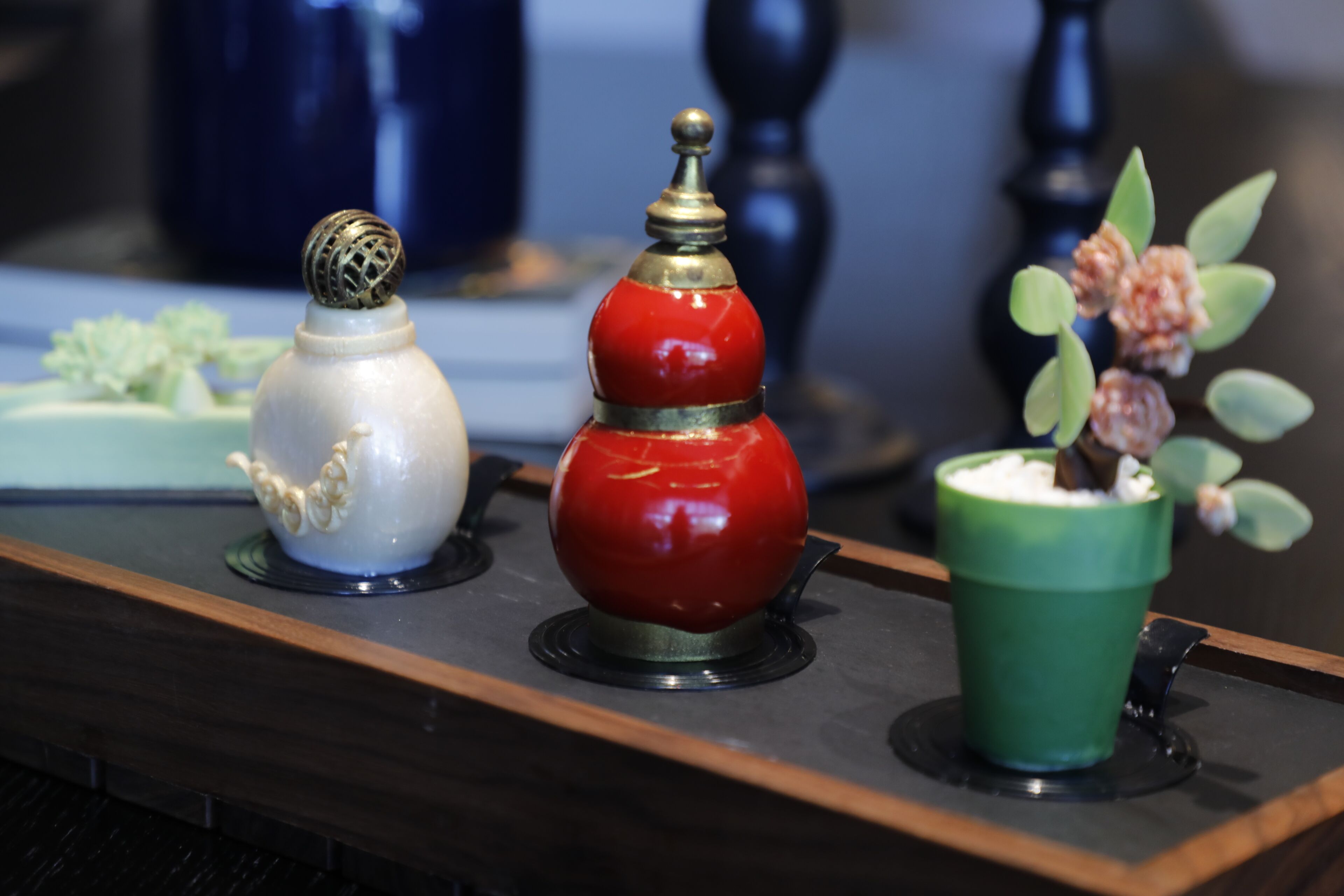 A tray with three decorative items: a white teapot, a red teapot, and a green cup with a plant.