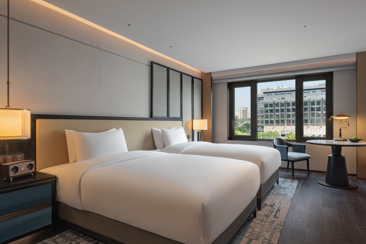 A hotel room with two beds, a desk, a chair, and a view of the cityscape through large windows.
