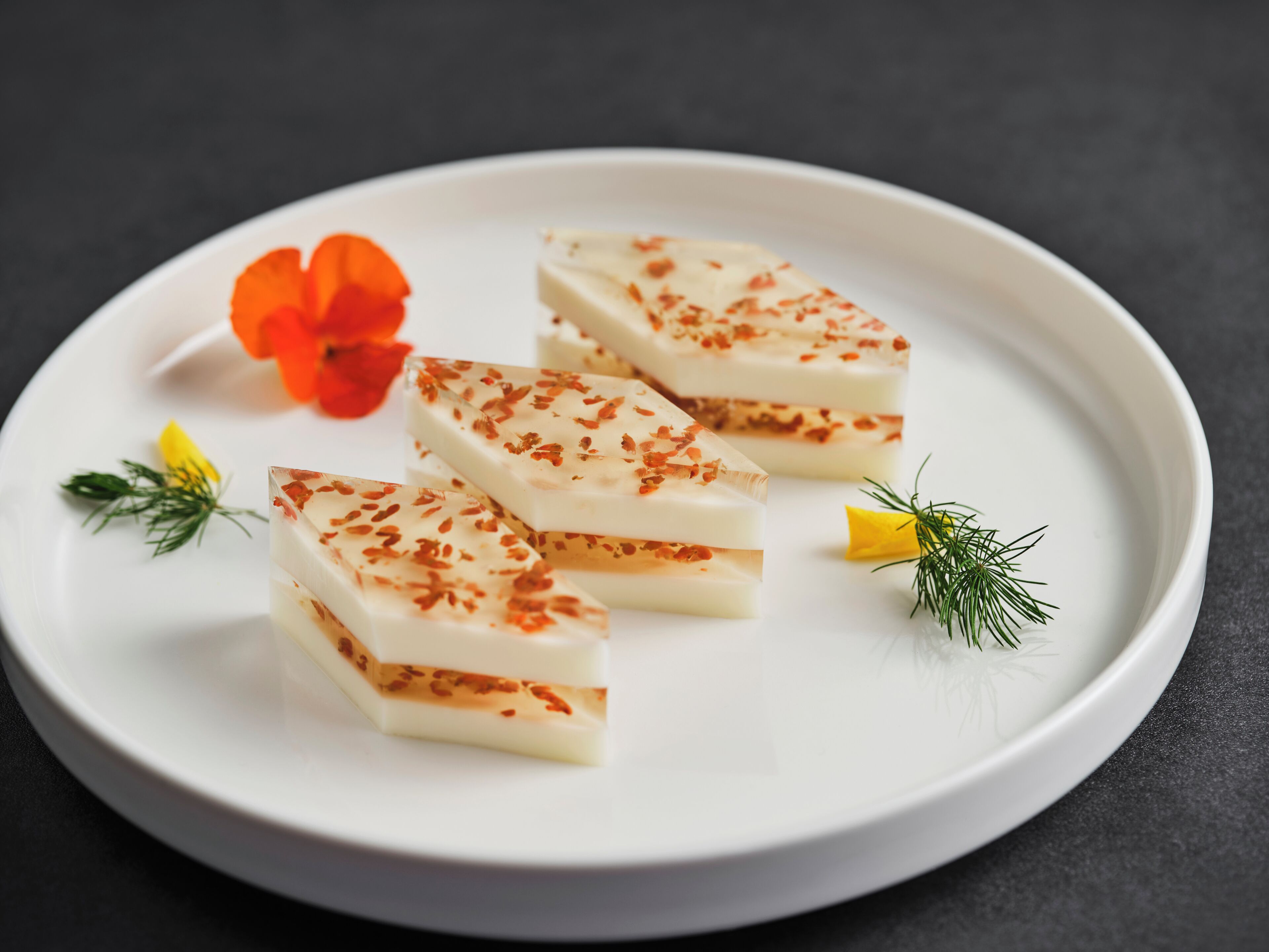 A white plate with rectangular food items garnished with red flakes, accompanied by a red flower and green herbs.