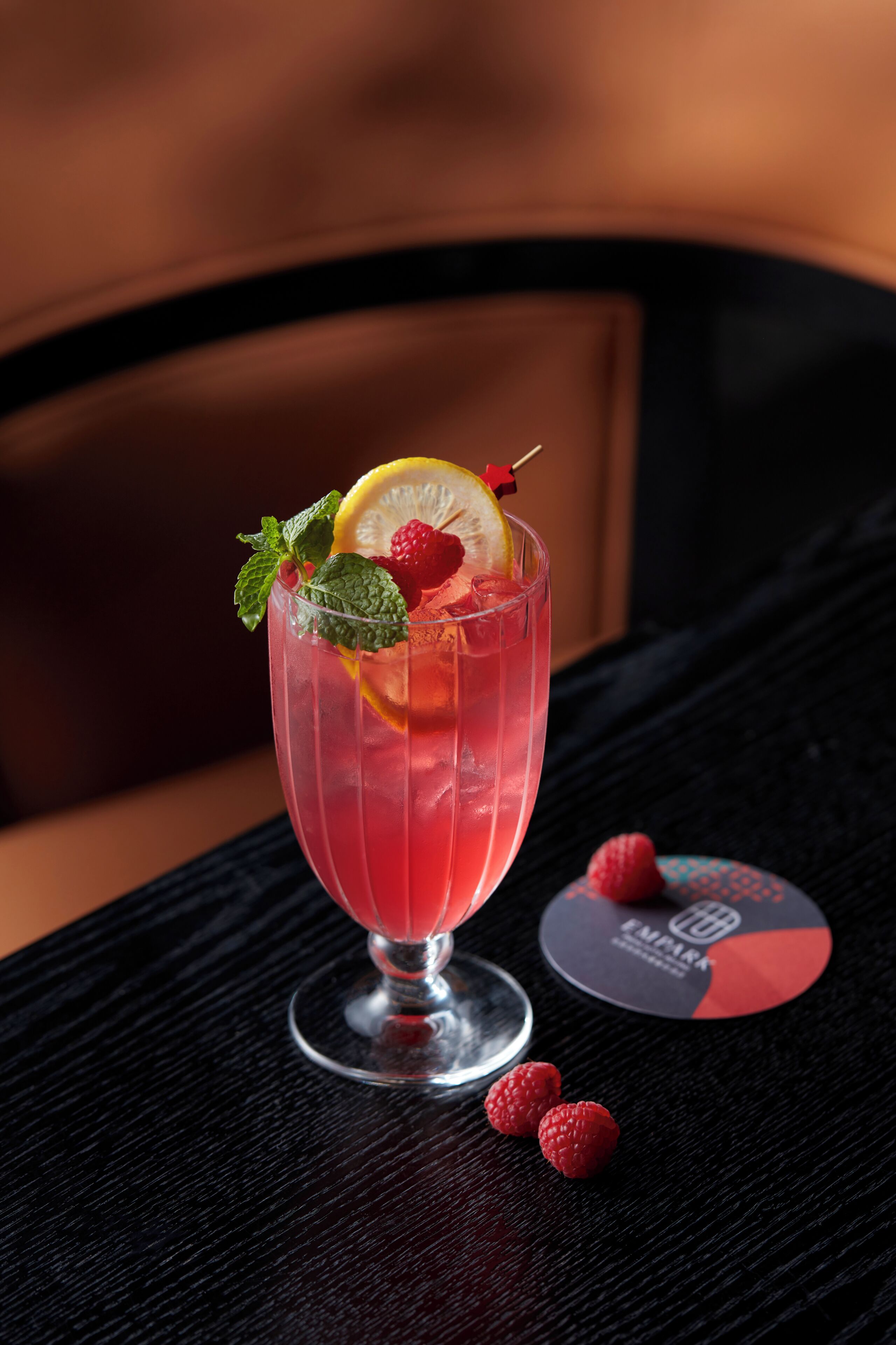 A cocktail glass with a slice of lemon, mint leaves, and raspberries on a black table.
