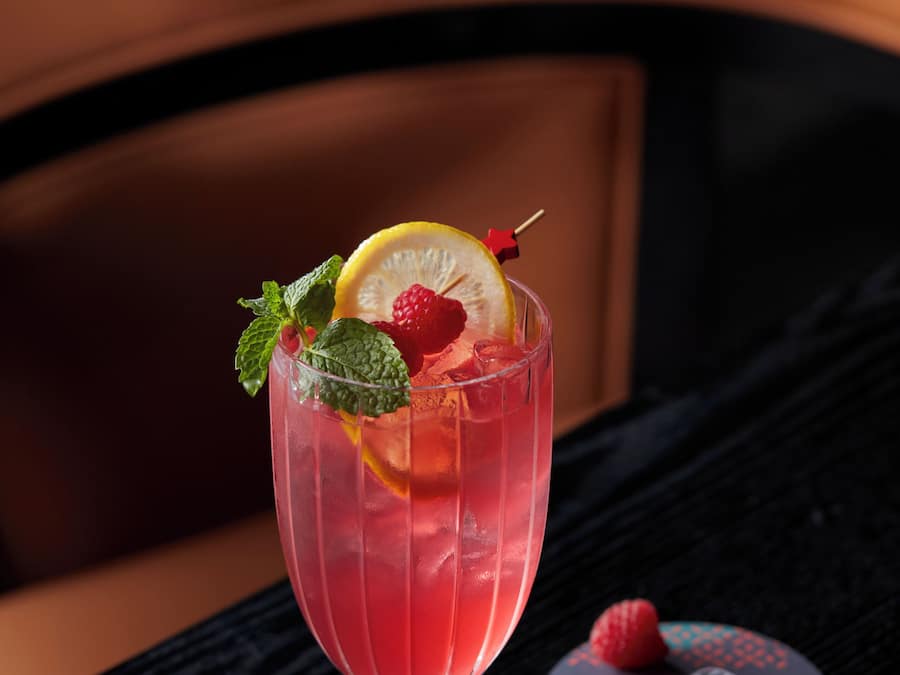 A cocktail glass with a slice of lemon, mint leaves, and raspberries on a black table.