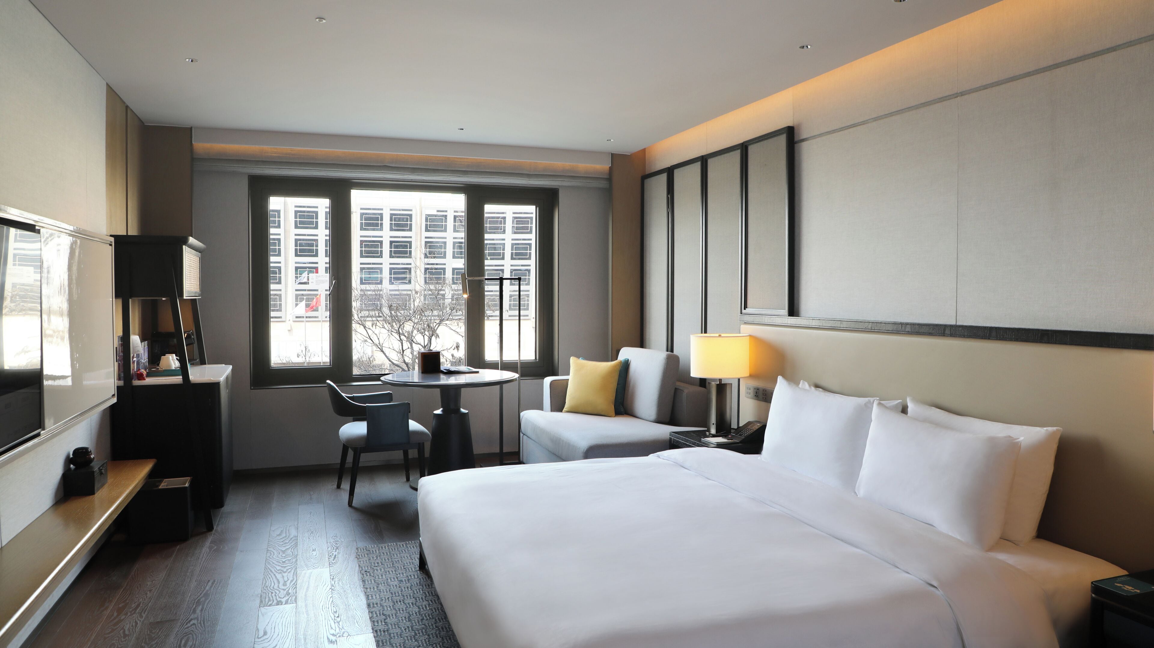 A hotel room with a bed, two chairs, a desk, a lamp, and a window with a view of buildings.