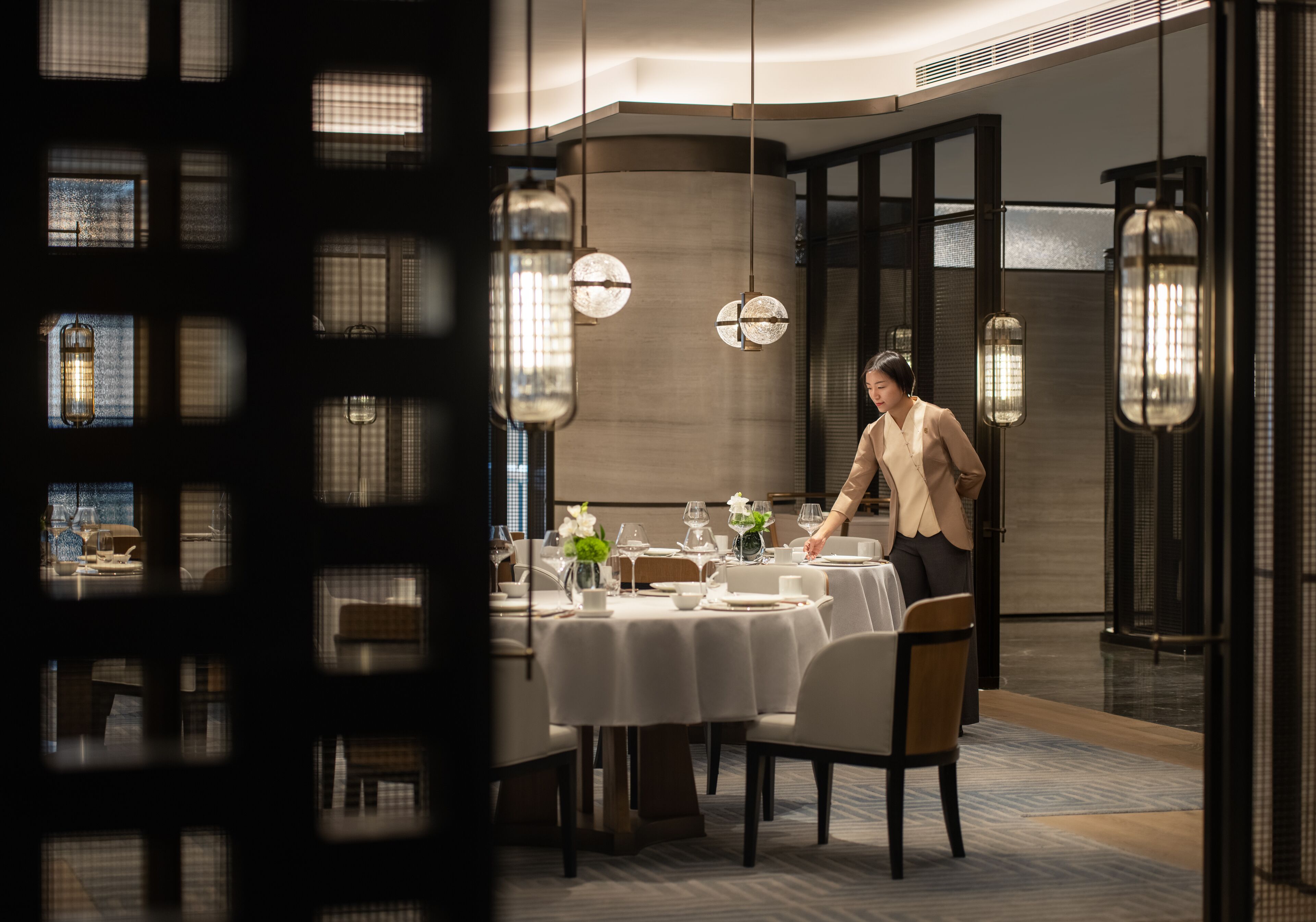 A dining area with round tables set for a meal, a person attending to the table, and hanging pendant lights.