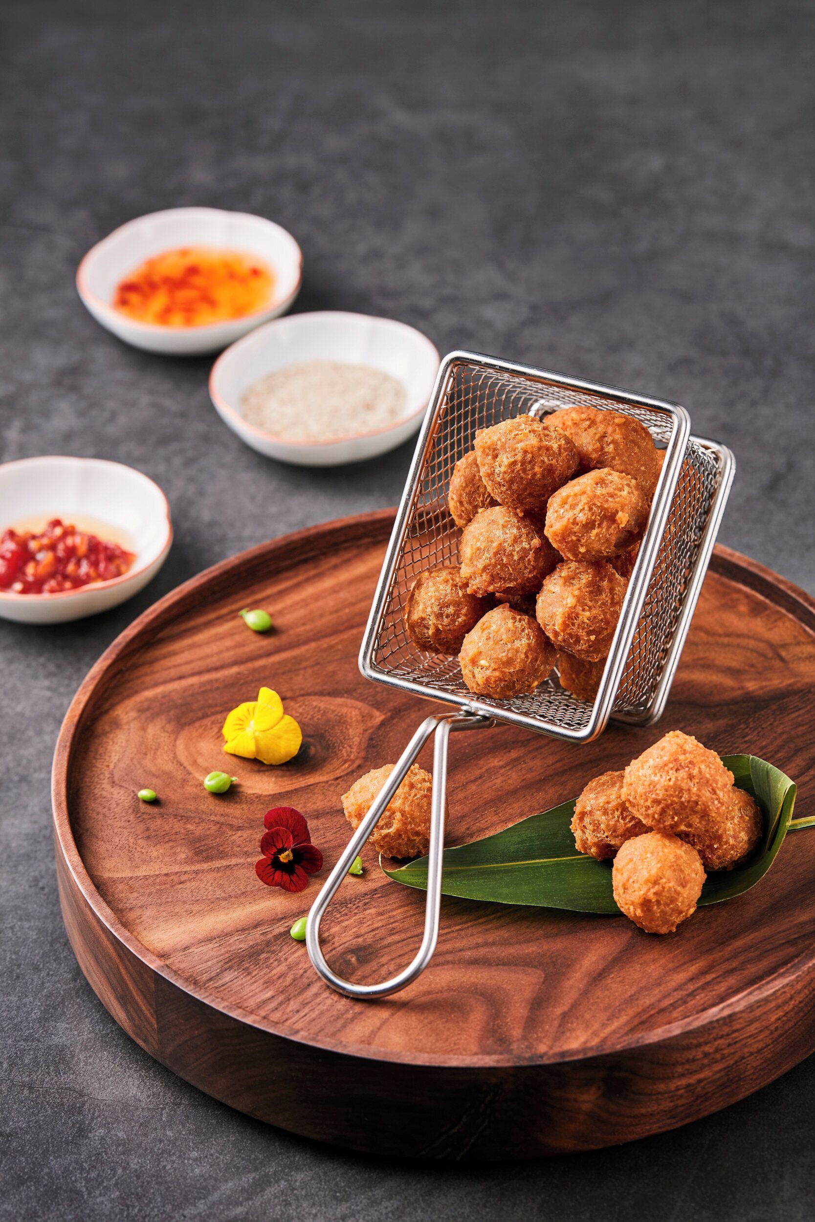 Una bandeja de madera con un colador de metal que contiene bolitas fritas, acompañada de recipientes pequeños con salsas y guarniciones.