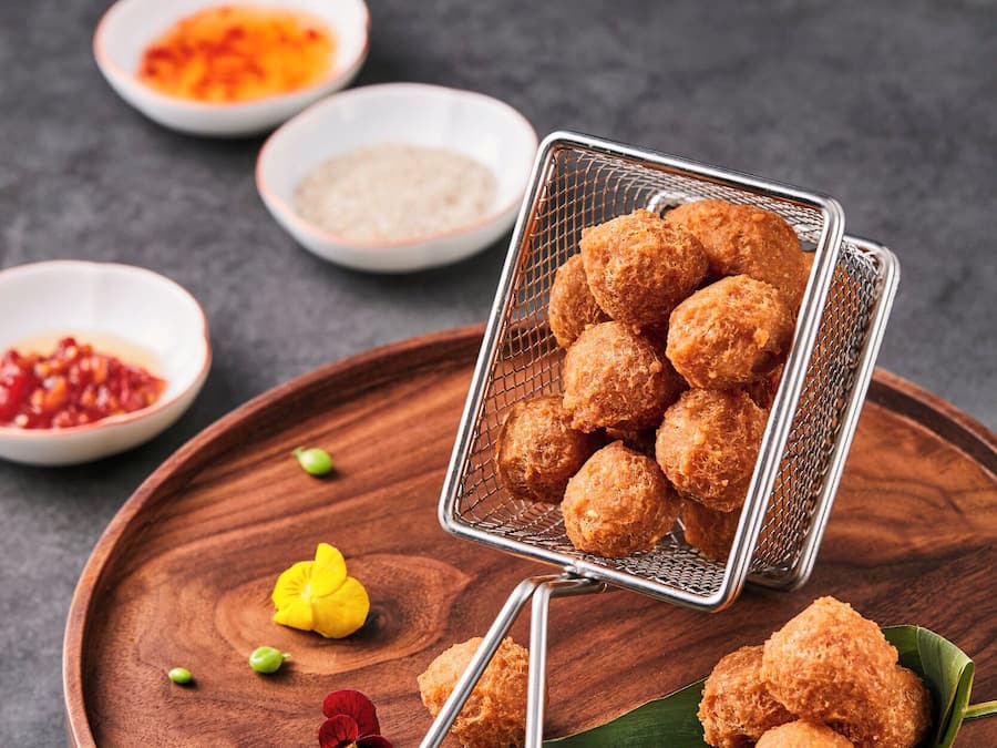 A wooden tray with a metal strainer containing fried balls, accompanied by small bowls of sauces and garnishes.