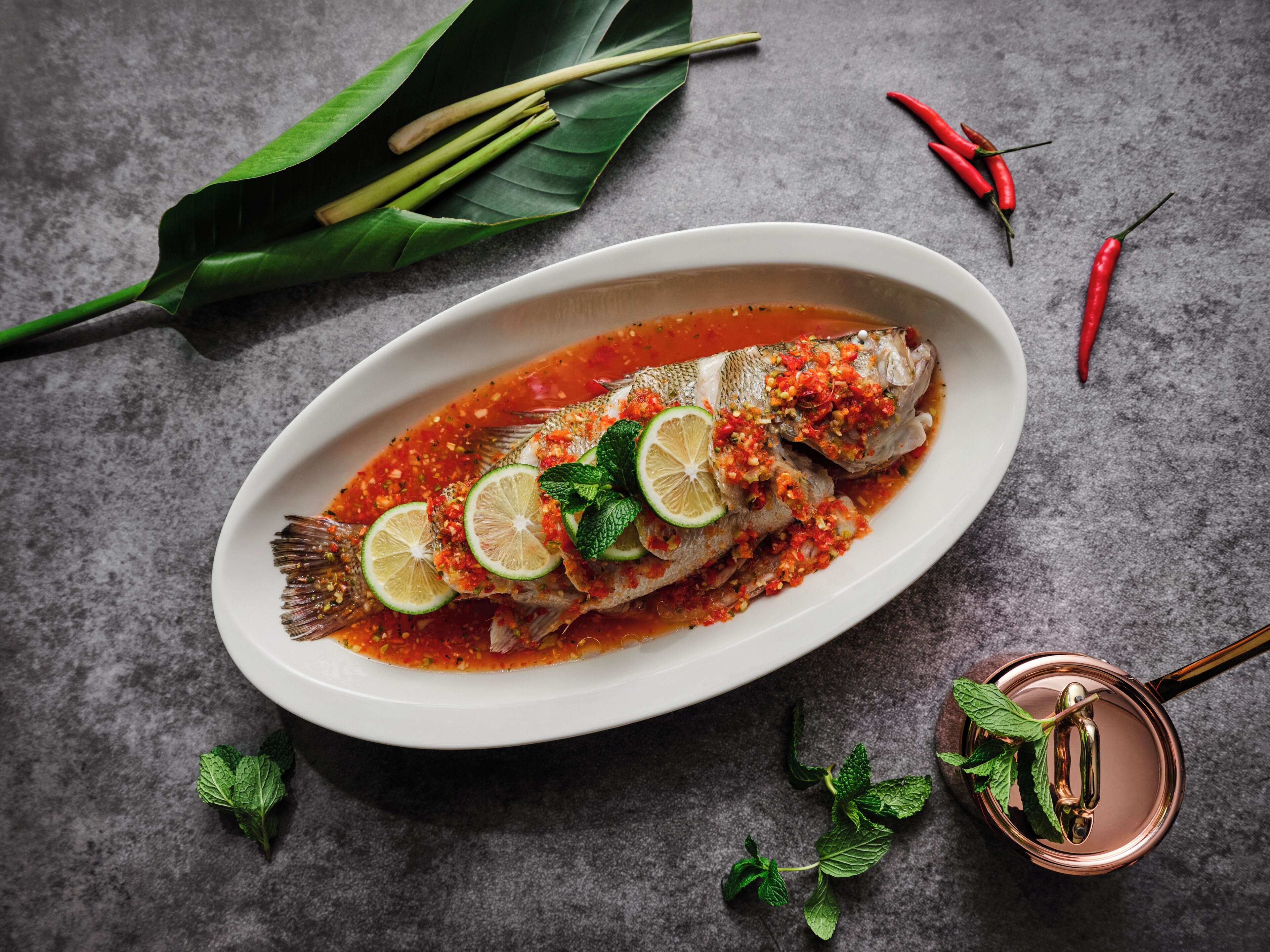 A white oval plate with a whole fish topped with lime slices and a red sauce, garnished with mint leaves, on a textured grey surface.