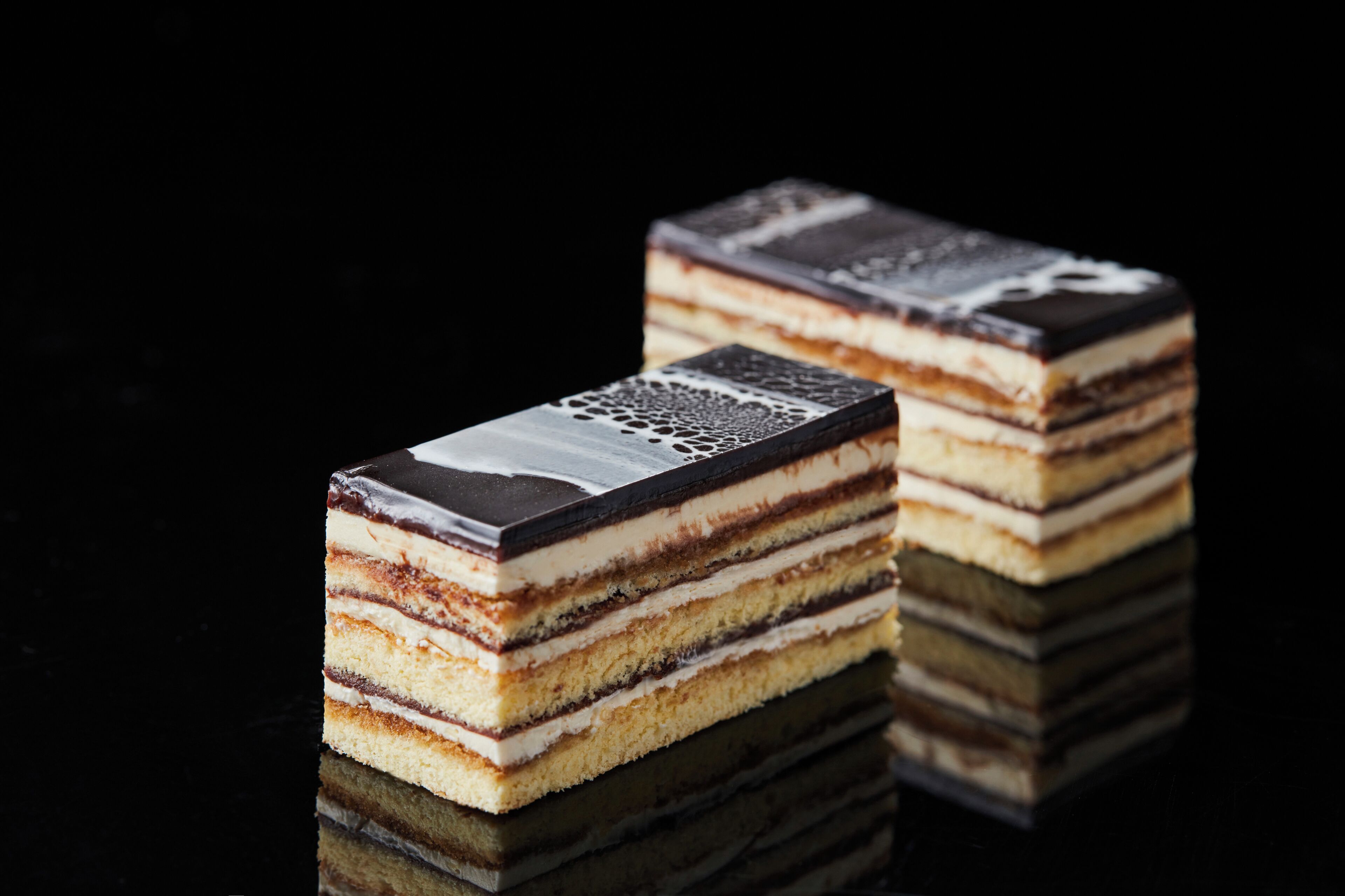 Two rectangular cakes with multiple layers and a decorative top, placed on a reflective surface.