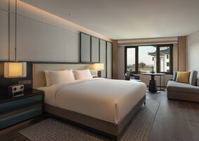 A modern hotel room with a large bed, bedside lamps, a small table with a chair, and a view of the city through large windows.