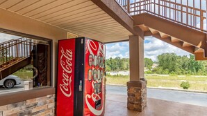 A Coca-Cola vending machine outside a building with a staircase and a view of a road and greenery.