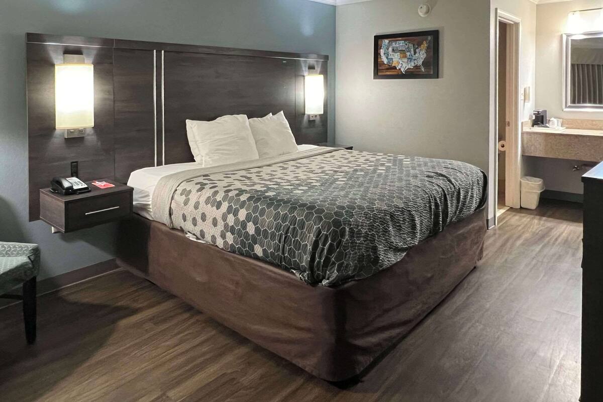 Habitación de hotel con una cama grande, dos lámparas de noche, un mesita de noche, una pequeña mesa y un baño visible a través de una puerta abierta.