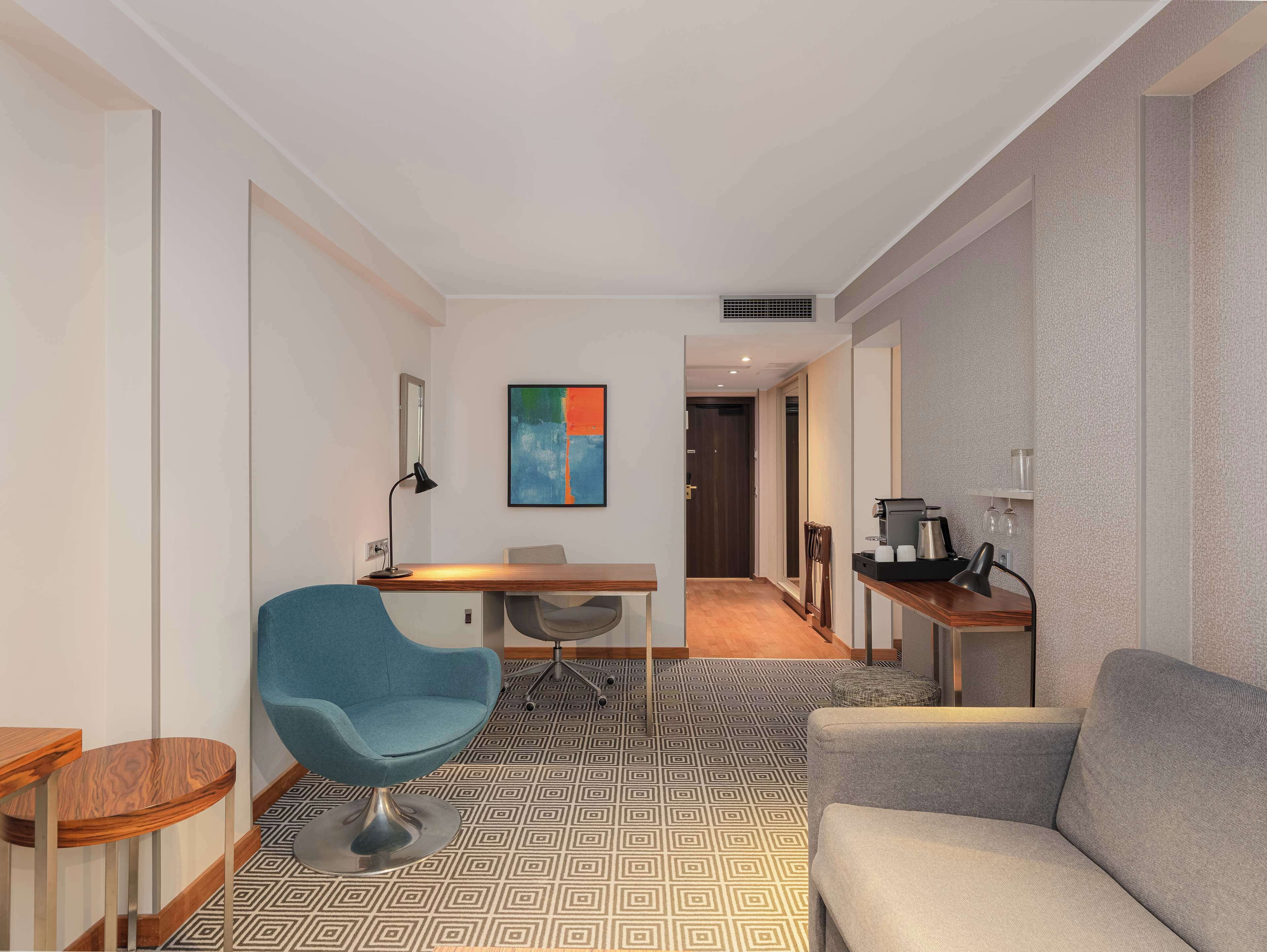 Une chambre d’hôtel moderne avec un fauteuil bleu, un canapé gris, une table basse en bois et un coin repas comprenant une petite table et des chaises.