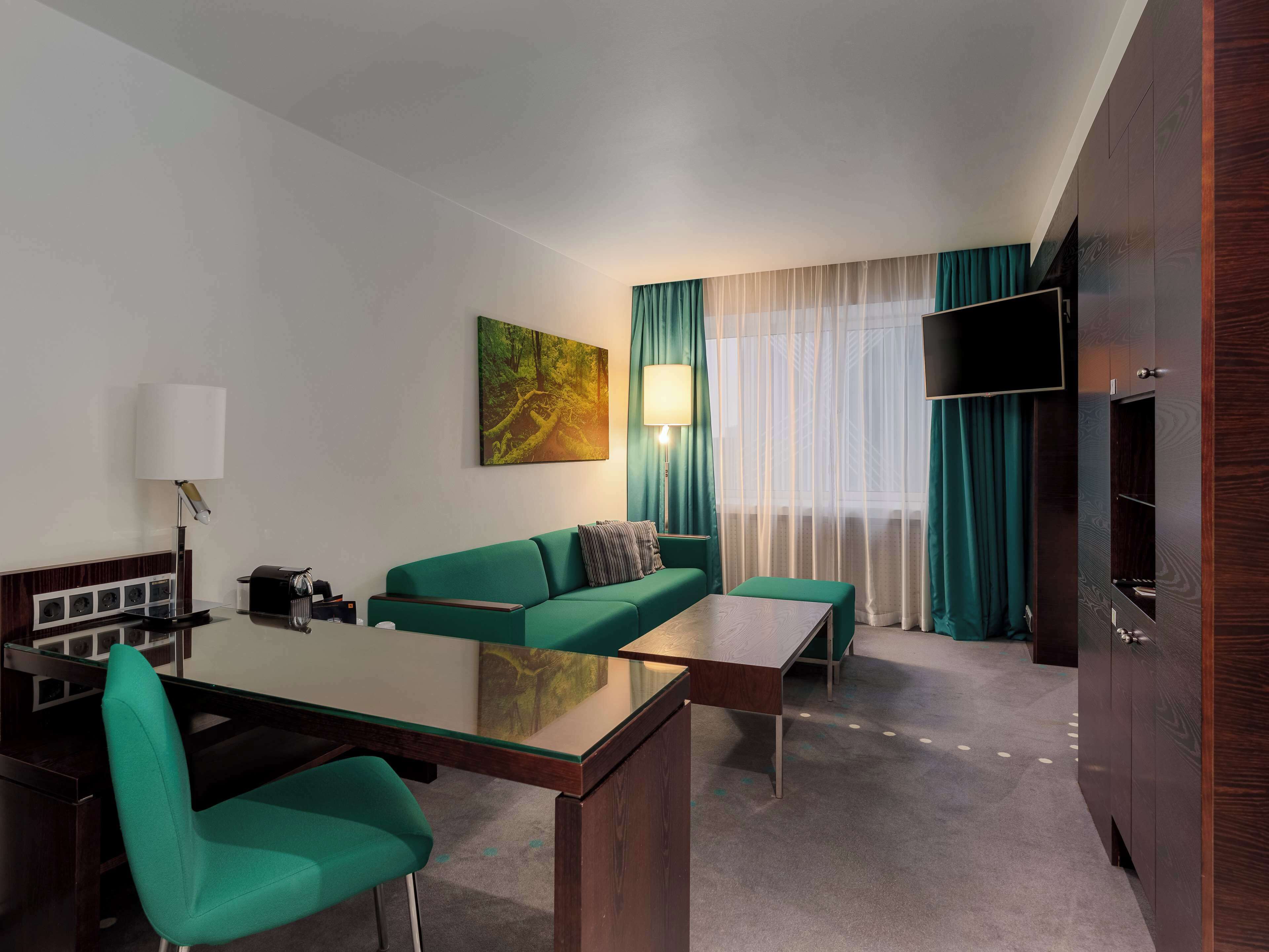 A modern hotel room with a green sofa, a glass dining table, a flat-screen TV, and a large window with sheer curtains.