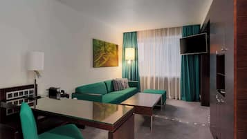 A modern hotel room with a green sofa, a glass dining table, a flat-screen TV, and a large window with sheer curtains.