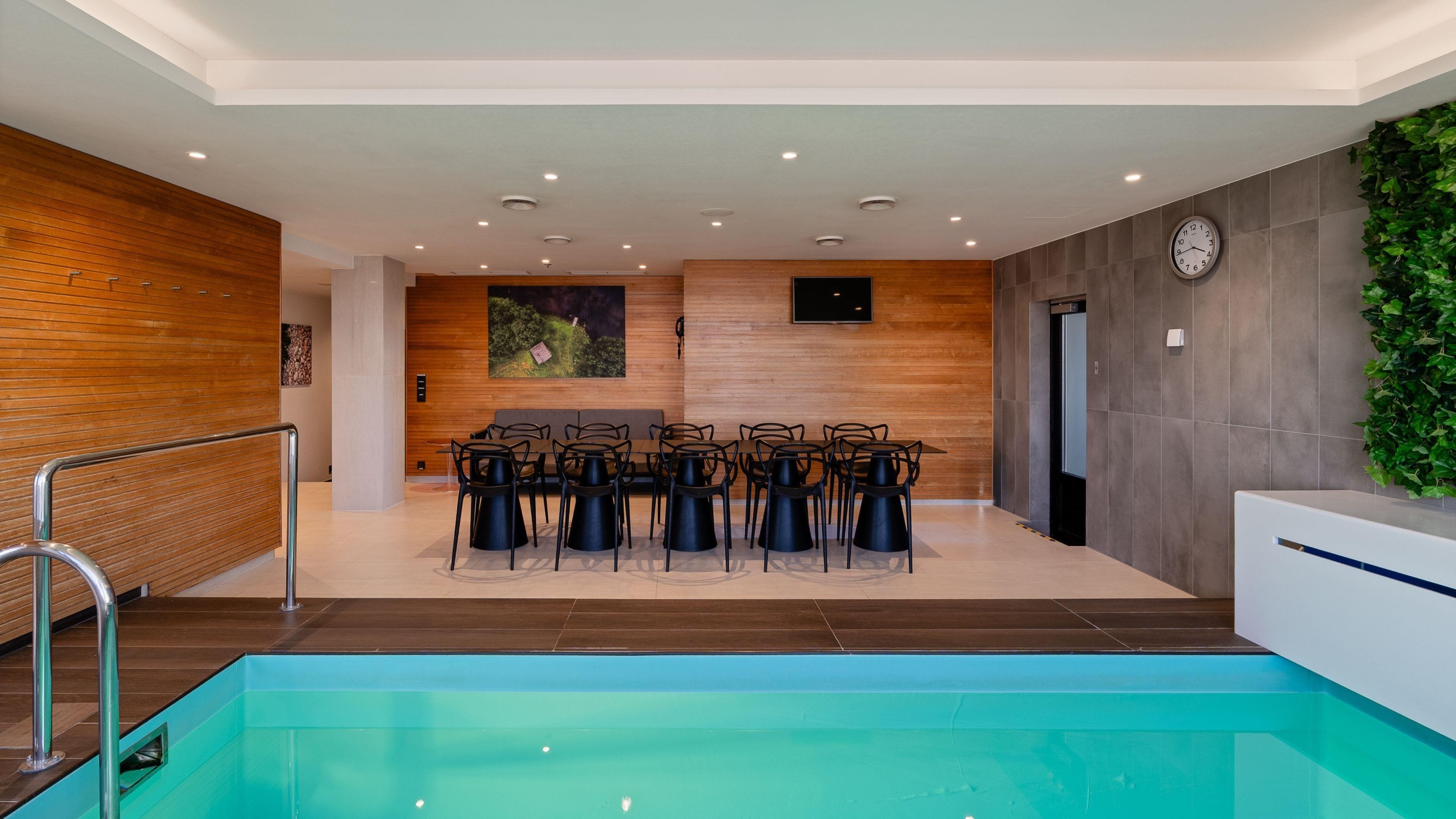 Un área interior de piscina moderna con una pared de madera, una zona de estar con sillas y un reloj en la pared.