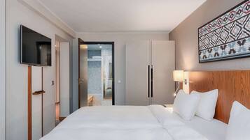 A hotel room with a large bed, a TV mounted on the wall, a wooden headboard, a bedside lamp, and a door leading to a bathroom.
