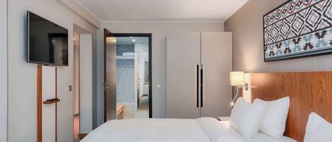 Habitación de hotel con una cama grande, televisor montado en la pared, cabecero de madera, lámpara de noche y puerta que da a un baño.