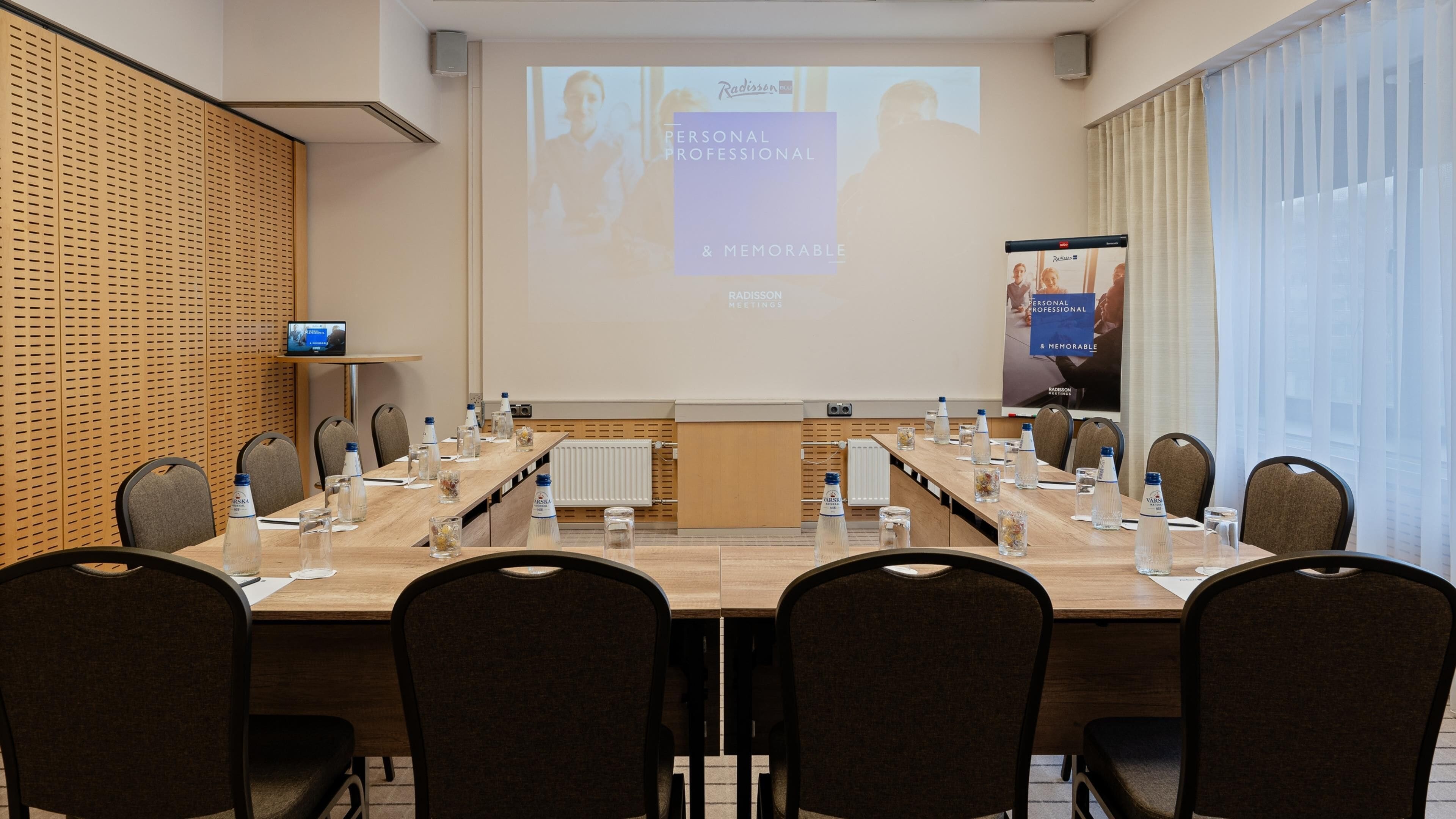 Een congrescentrum met een lange tafel, stoelen, een projectiescherm en een scherm waarop 'Radisson Meetings' staat.