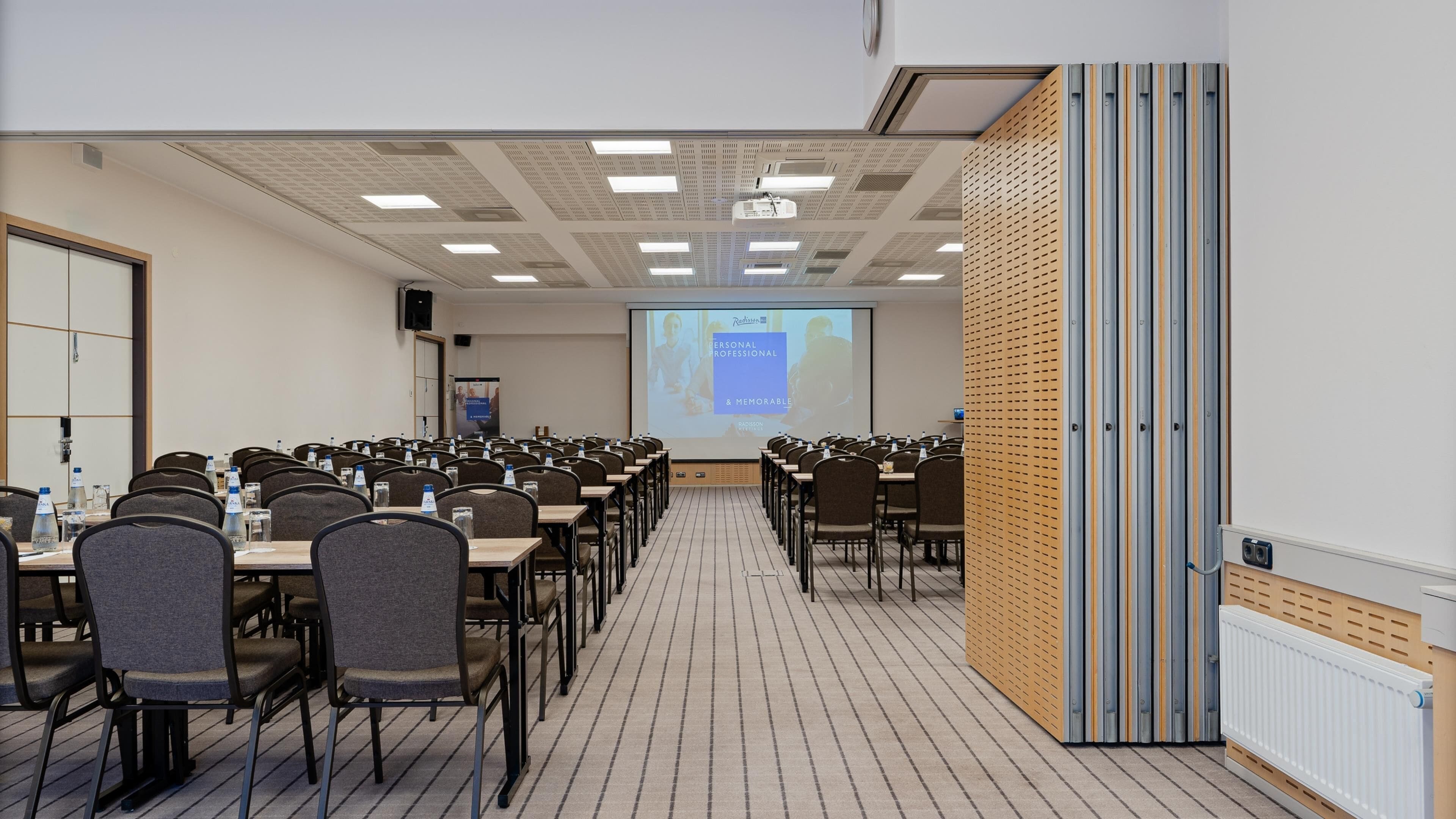 Een vergaderruimte met rijen tafels en stoelen, een projectiescherm waarop een presentatie wordt getoond en een speakersysteem.