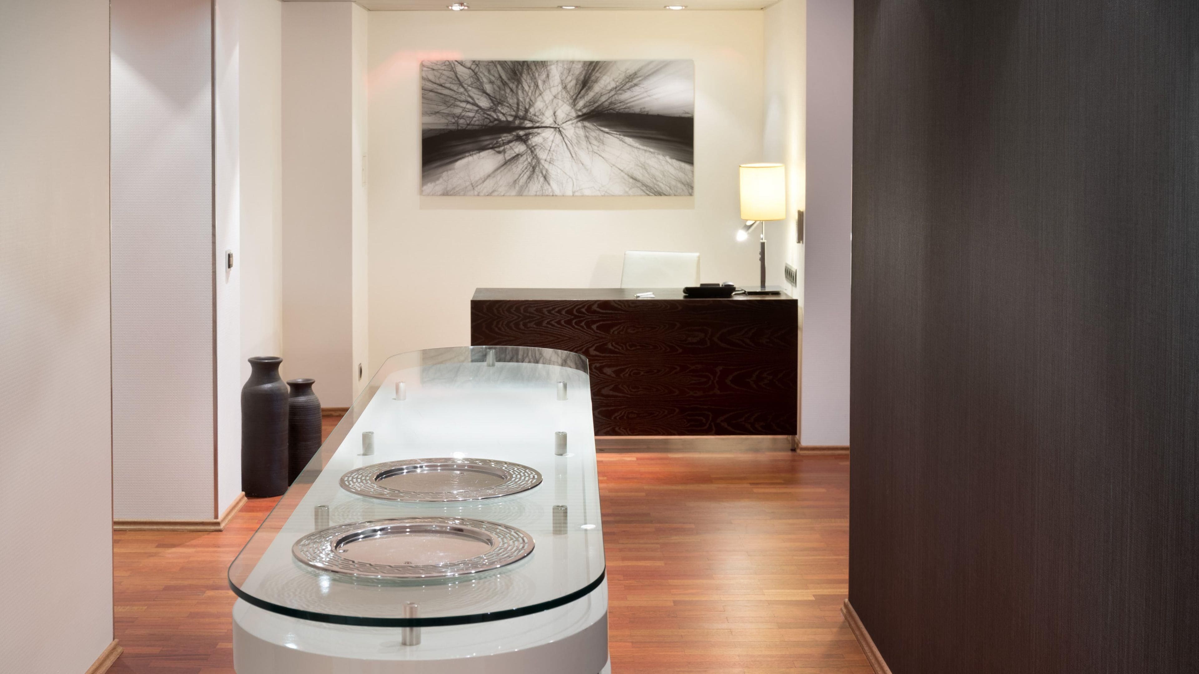 Zona de recepción con una mesa de vidrio, suelo de madera, un mostrador oscuro y un cuadro abstracto colgado en la pared.