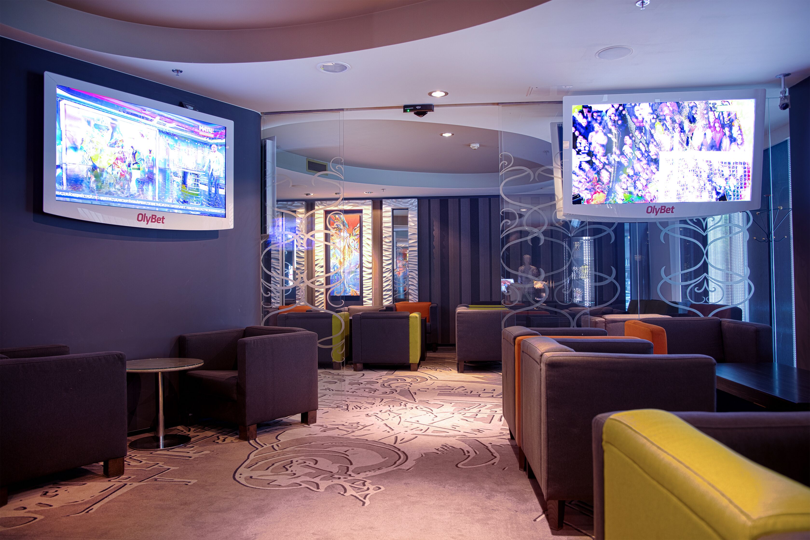 A modern lounge area with comfortable seating, a patterned carpet, and two large screens displaying content.