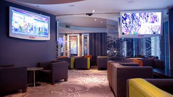 A modern lounge area with comfortable seating, a patterned carpet, and two large screens displaying content.