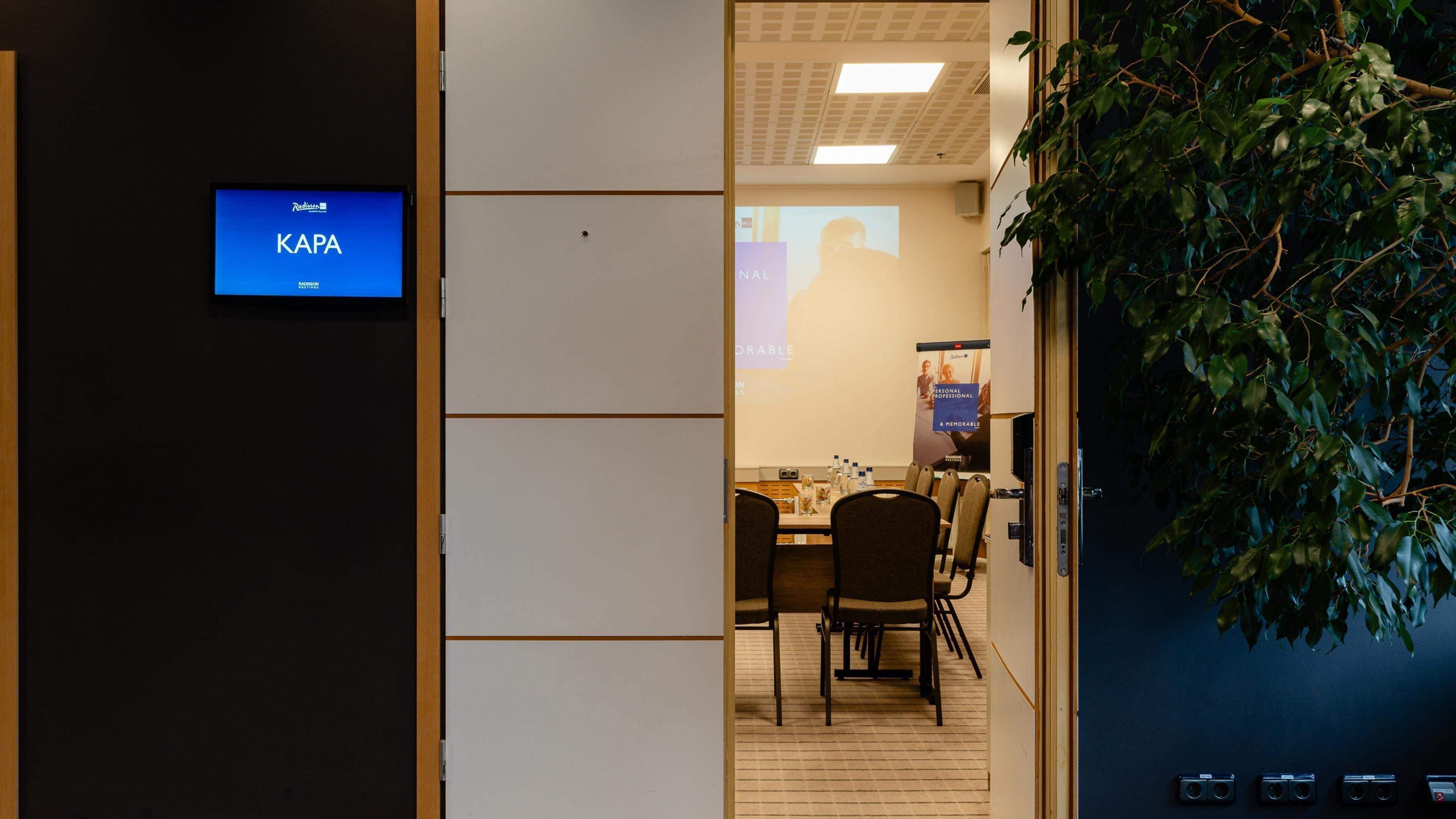 A room with a KAPA sign, a projector screen, a table with chairs, and a plant.