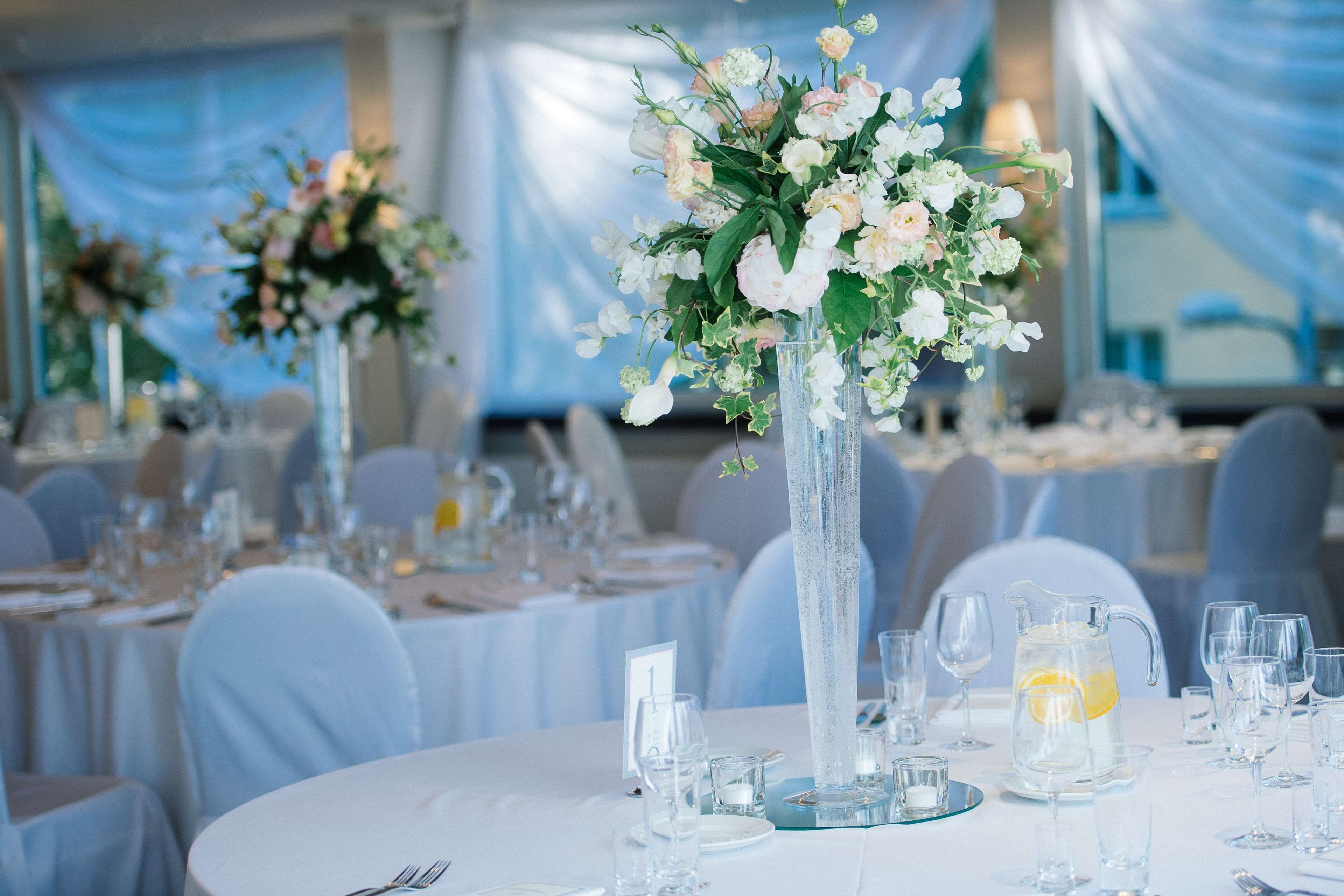 Une salle de banquet avec des tables rondes dressées pour un événement formel, agrémentée de grands arrangements floraux et de verres transparents.