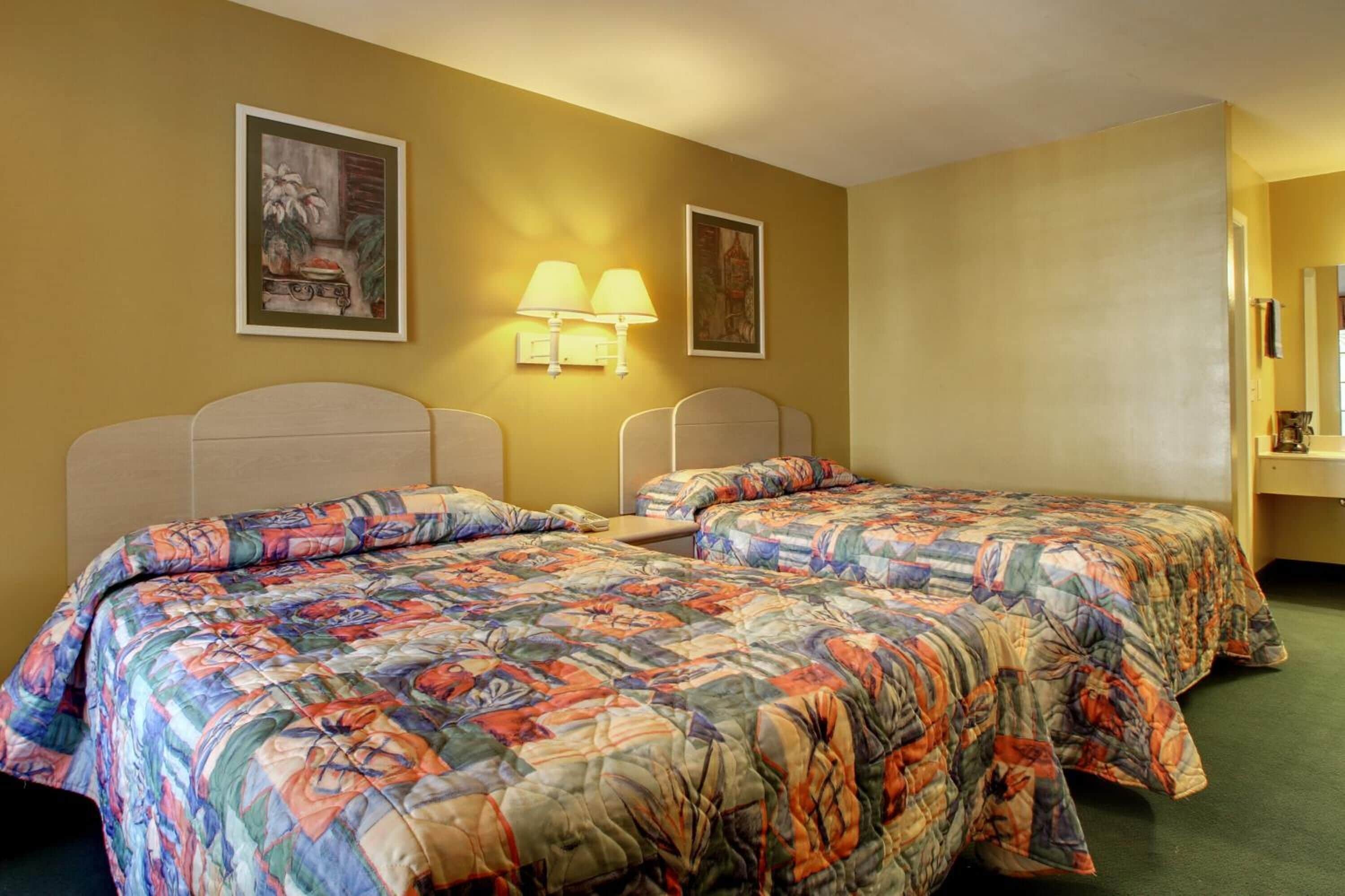Habitación de hotel con dos camas, cada una con un edredón colorido, una mesita de noche, una lámpara de pared y dos cuadros enmarcados.