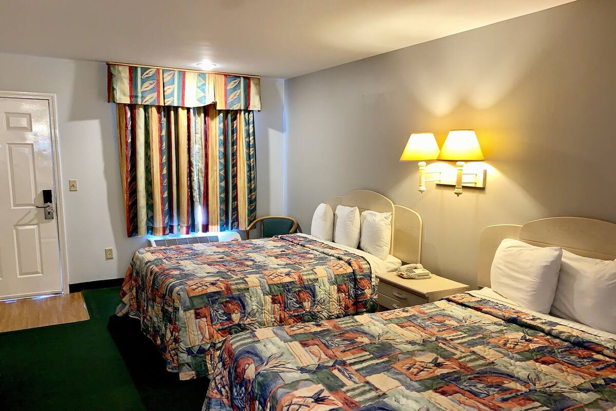 A hotel room with two beds, a window with colorful curtains, a bedside lamp, and a green carpet.