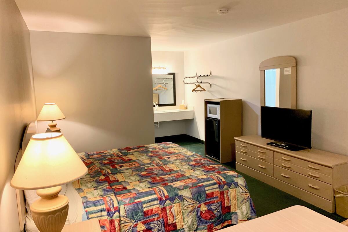 A hotel room with a bed, a dresser, a television, a desk, a lamp, a mirror, and a small kitchenette.
