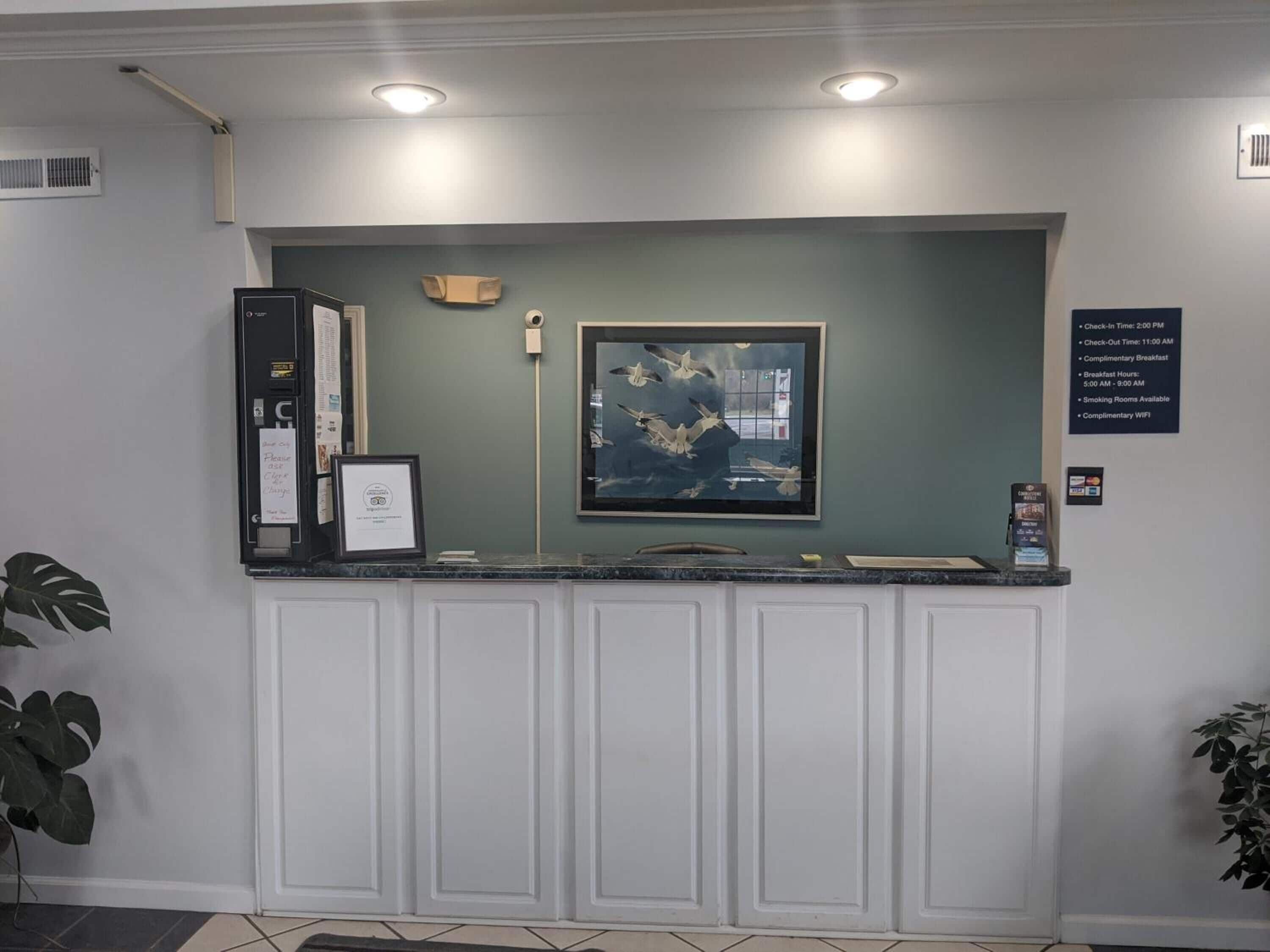 A reception desk with a framed artwork, a check-in/out board, and a sign detailing amenities like complimentary breakfast and WiFi.