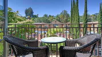 A balcony with wicker chairs and a table, overlooking a courtyard with buildings and tall trees.