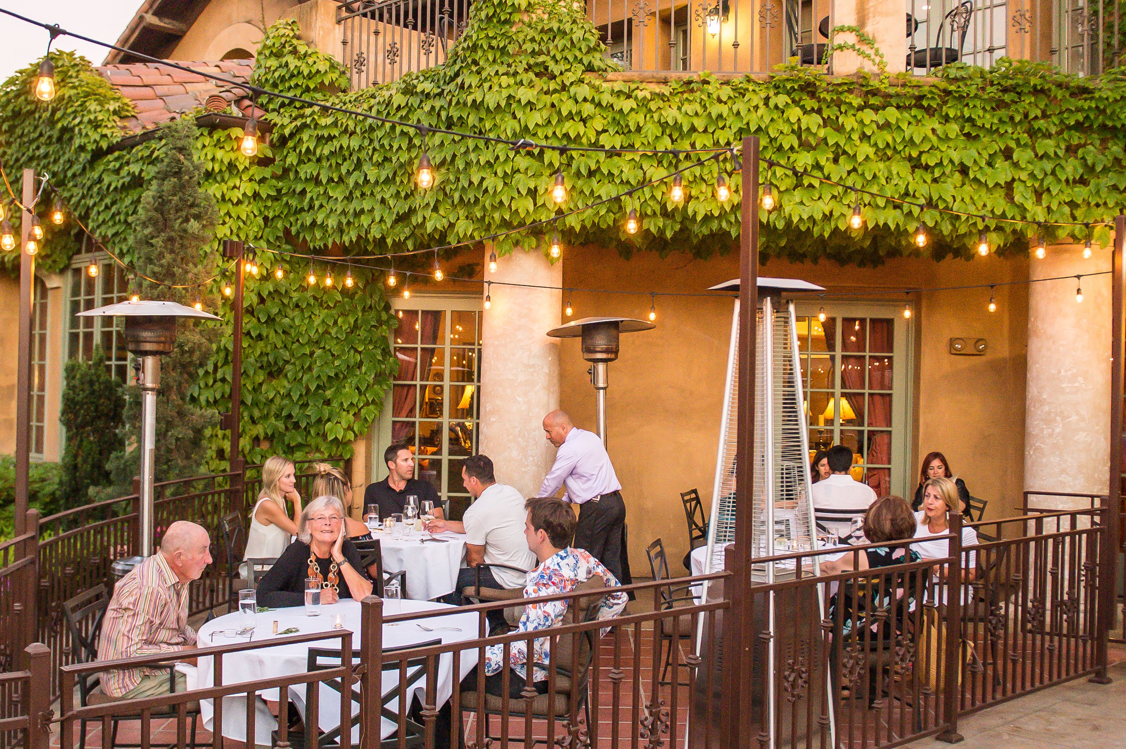 An outdoor dining area with tables and chairs, covered by a green vine.