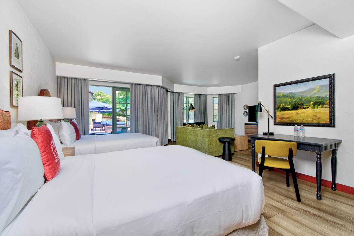 A hotel room with two beds, a desk, a chair, a television, and a view of the outdoors.