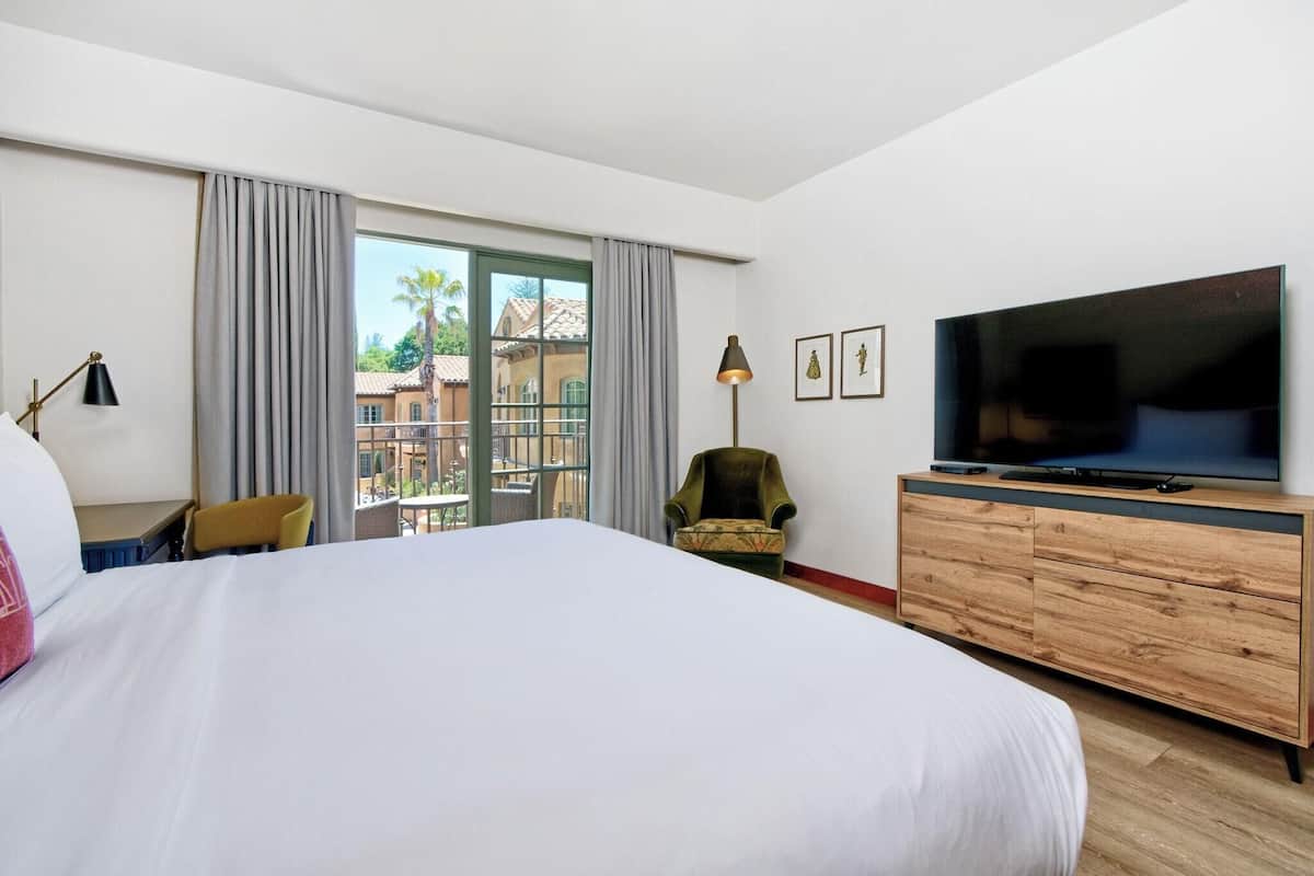 A hotel room with a large bed, a television, a wooden dresser, a chair, and a balcony with a view of buildings and palm trees.