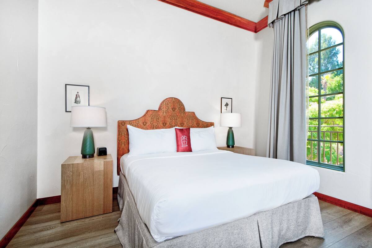 A neatly made bed with white linens, two bedside tables with lamps, and a window with a view of greenery.