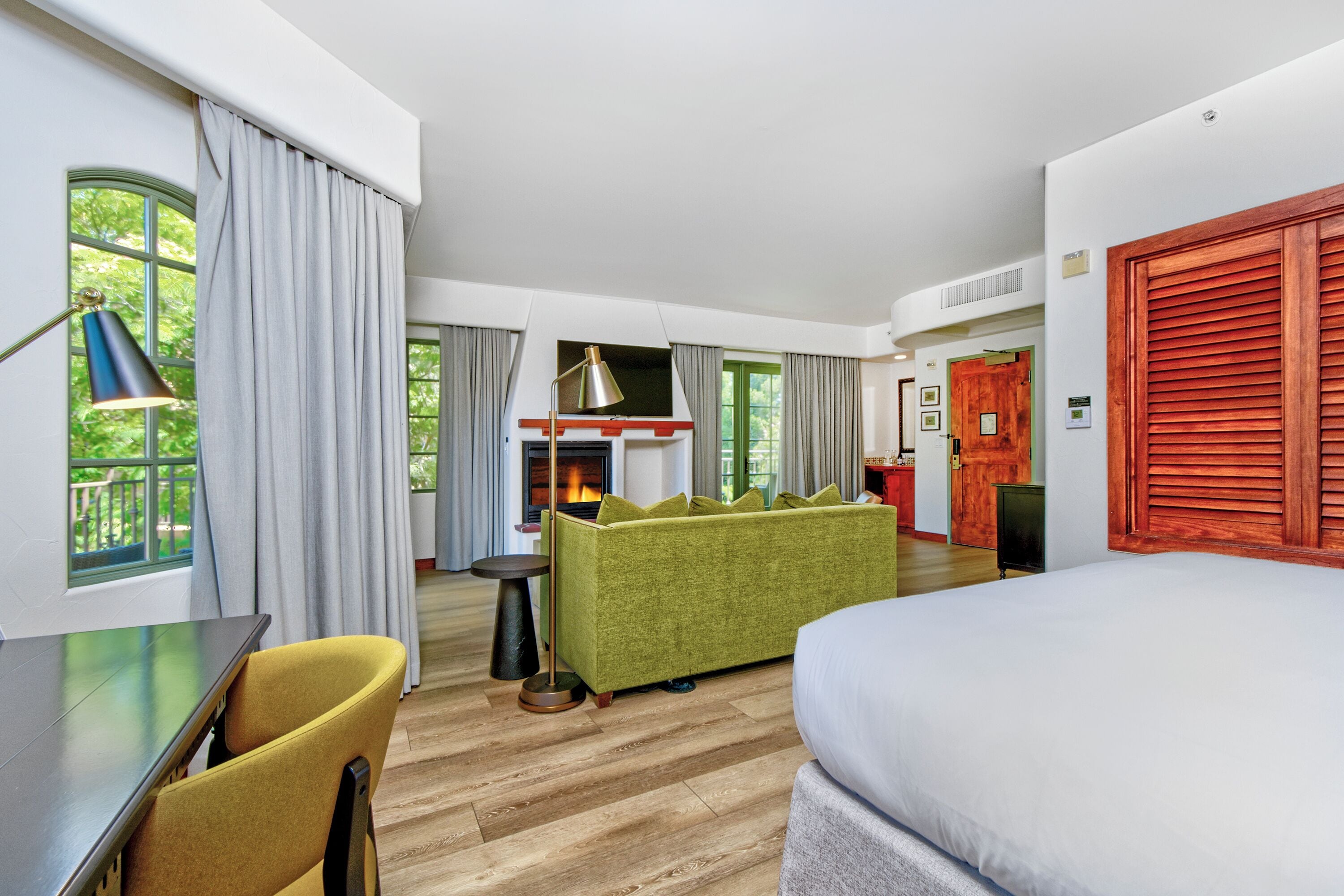Habitación de hotel con cama, sofá, mesa de comedor con sillas, chimenea y vistas a la vegetación.