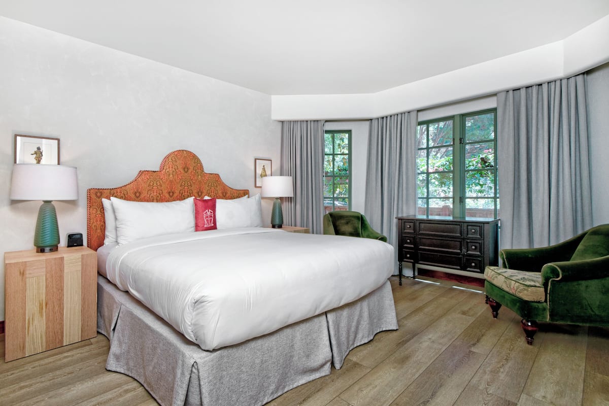 A hotel room with a large bed, two bedside tables, a dresser, a green armchair, and a window with curtains.