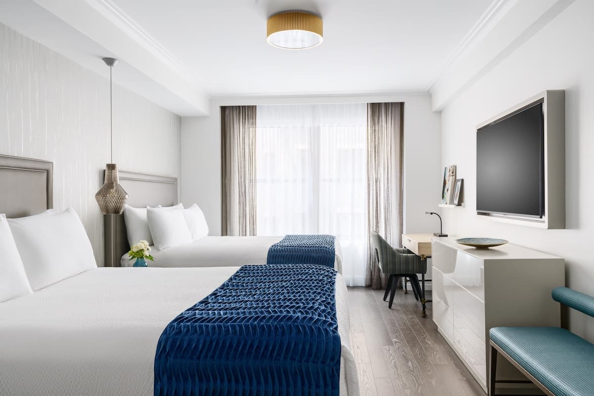 Habitación de hotel con dos camas, un escritorio, un televisor y un amplio ventanal con cortinas.