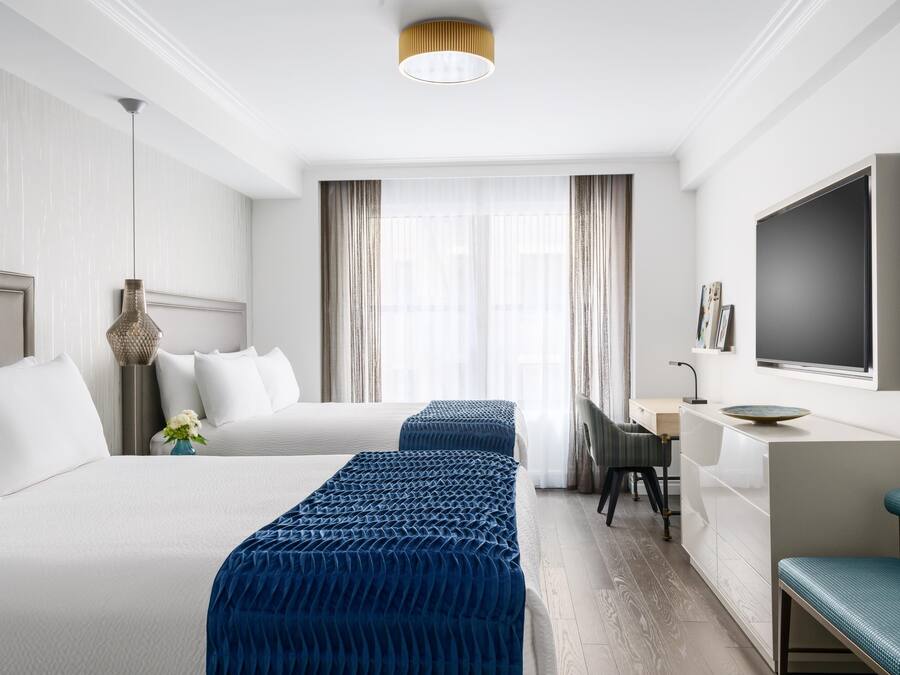 A hotel room with two beds, a desk, a TV, and a large window with curtains.