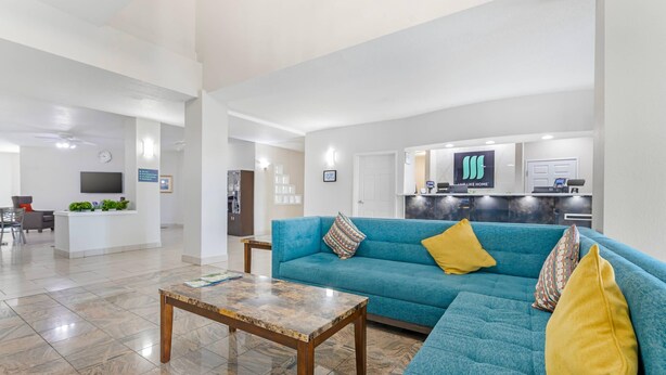 A modern living room with a teal sofa, wooden coffee table, and yellow cushions. A reception desk with a TV and a sign reading "LAPLIKE HOME" is in the background. - MainStay Suites Madison Airport (Madison)