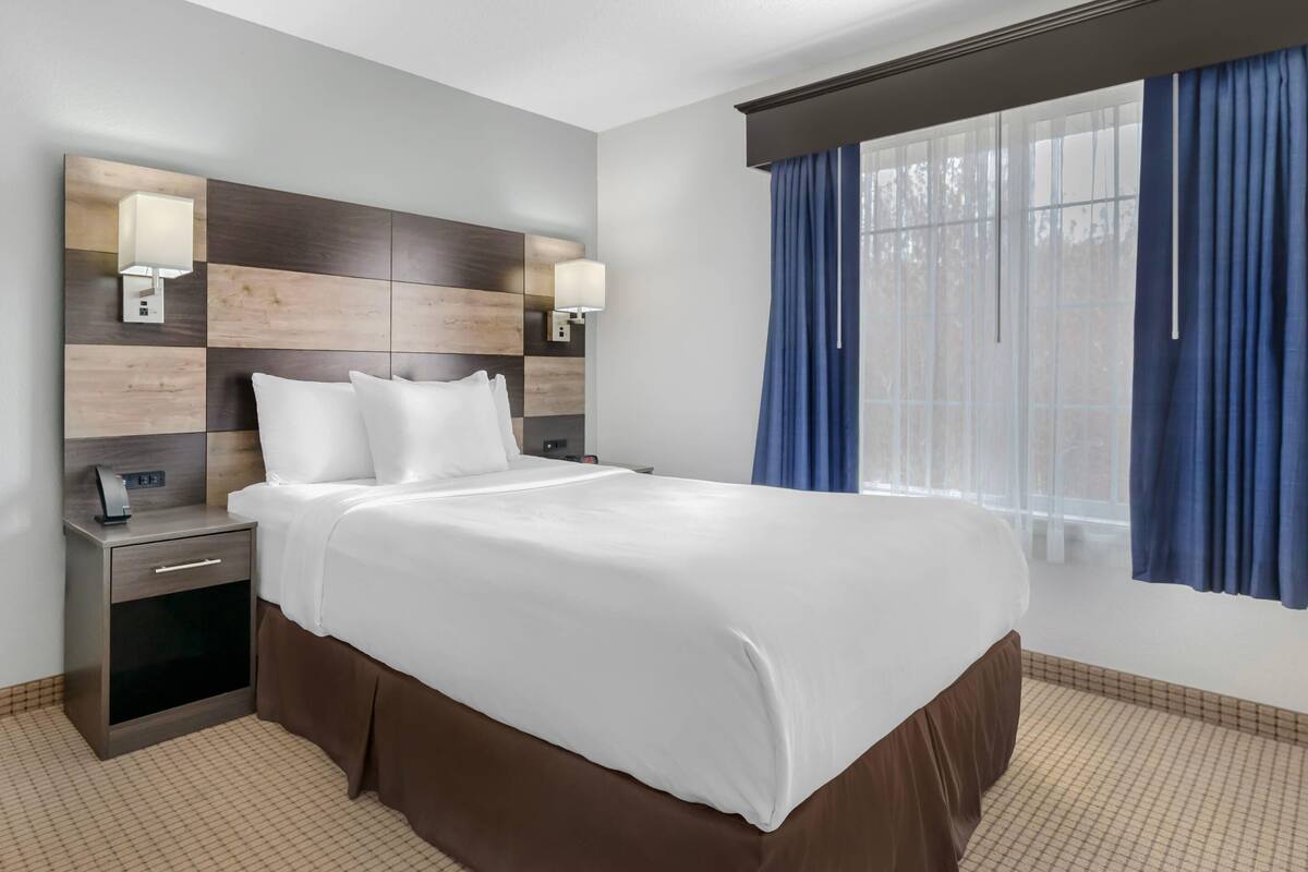 Une chambre d’hôtel avec un grand lit, une table de chevet, une lampe fixée au mur et une fenêtre avec des rideaux.
