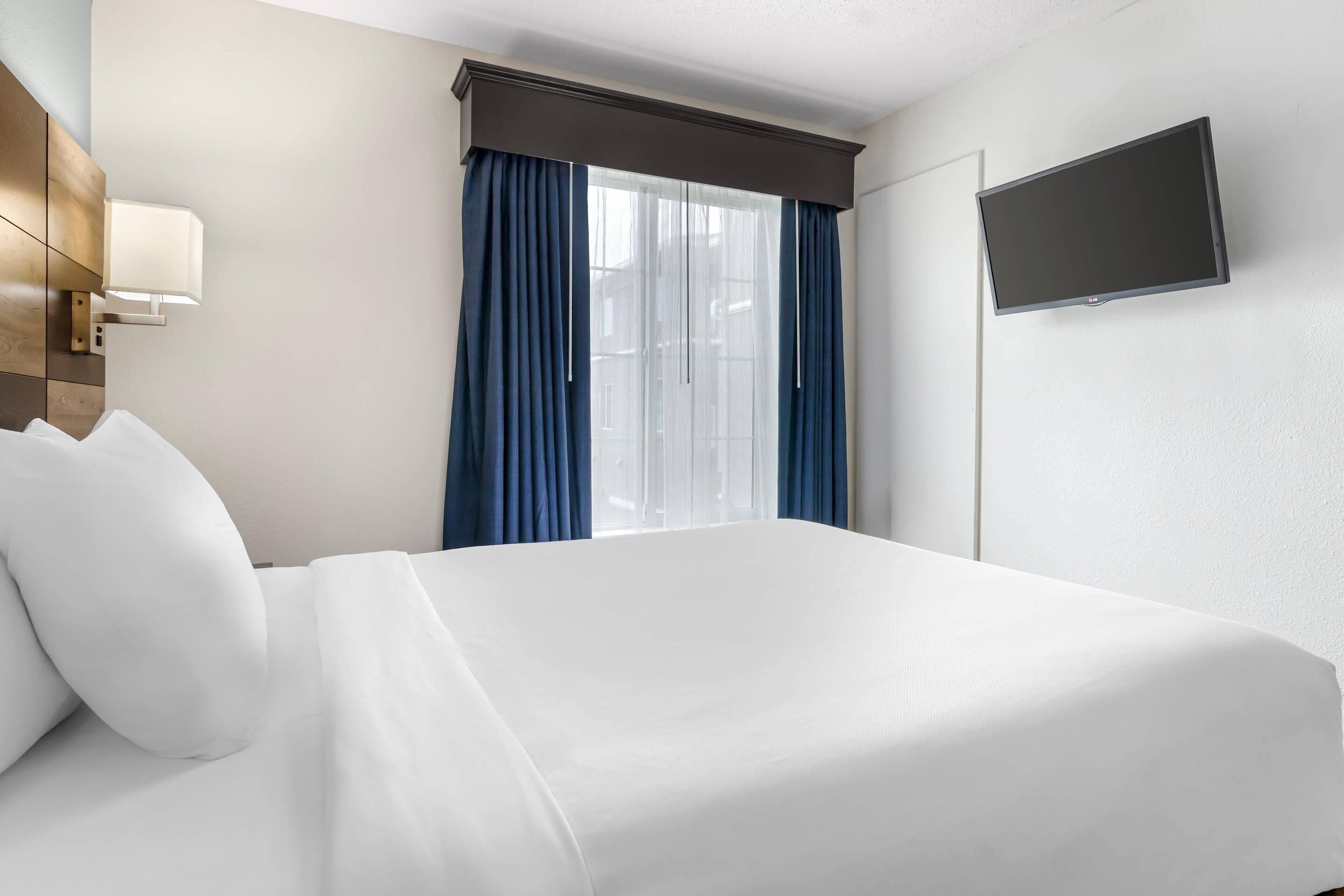 A hotel room with a bed, a flat-screen TV mounted on the wall, and a window with blue curtains.