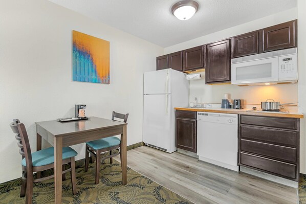 A compact kitchen with dark wooden cabinets, a white refrigerator, a microwave, and a dishwasher. There is a dining table with two chairs and a small rug on the floor. - MainStay Suites Madison Airport (Madison)