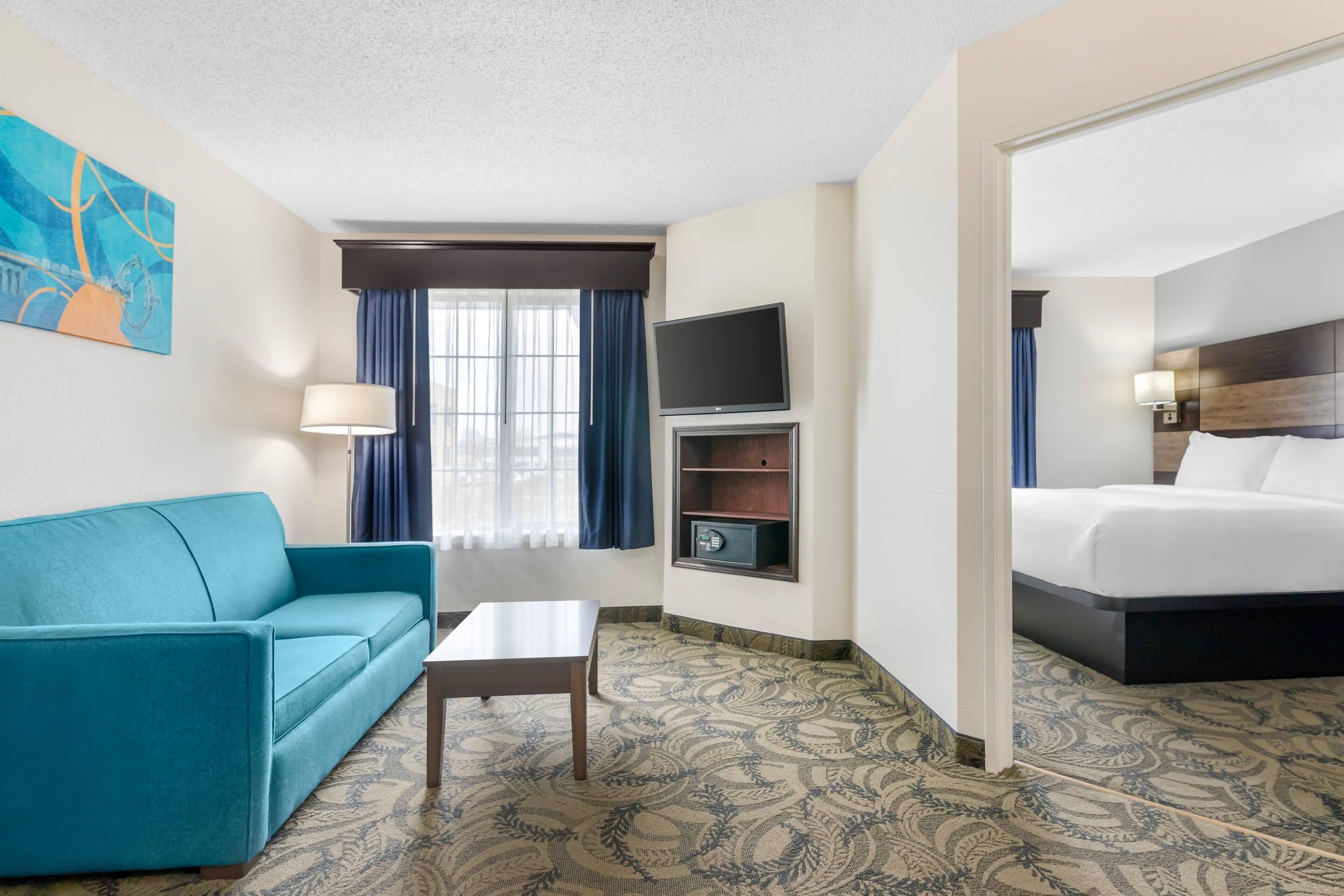 A hotel room with a teal sofa, a small table, a TV, a bed, and a patterned carpet.